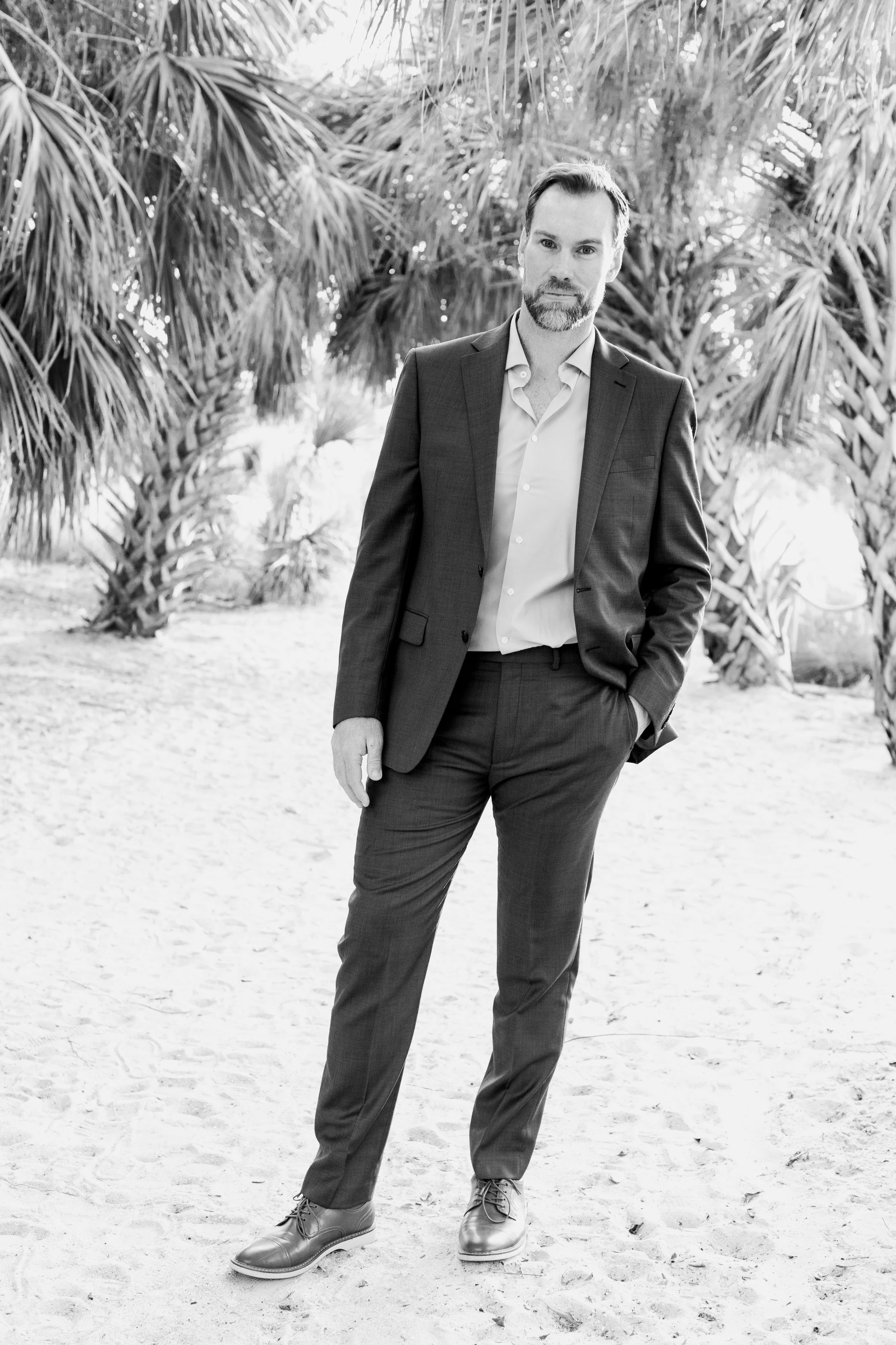 A man in a dark suit and light-colored shirt standing outdoors on sandy ground with trees and palm fronds in the background.