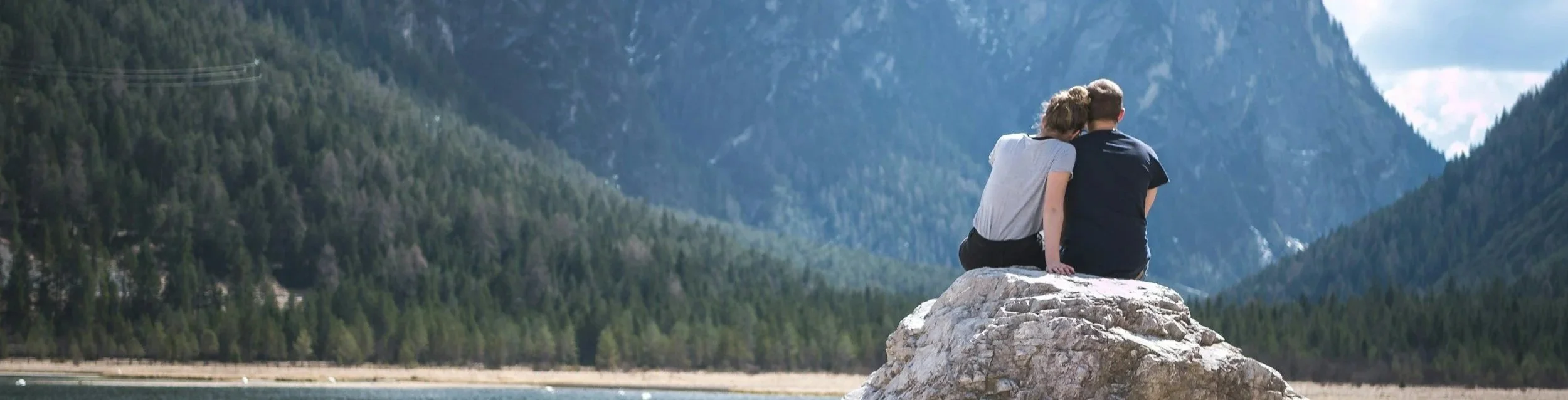 Two people sitting on a large rock watching the mountains and forested landscape