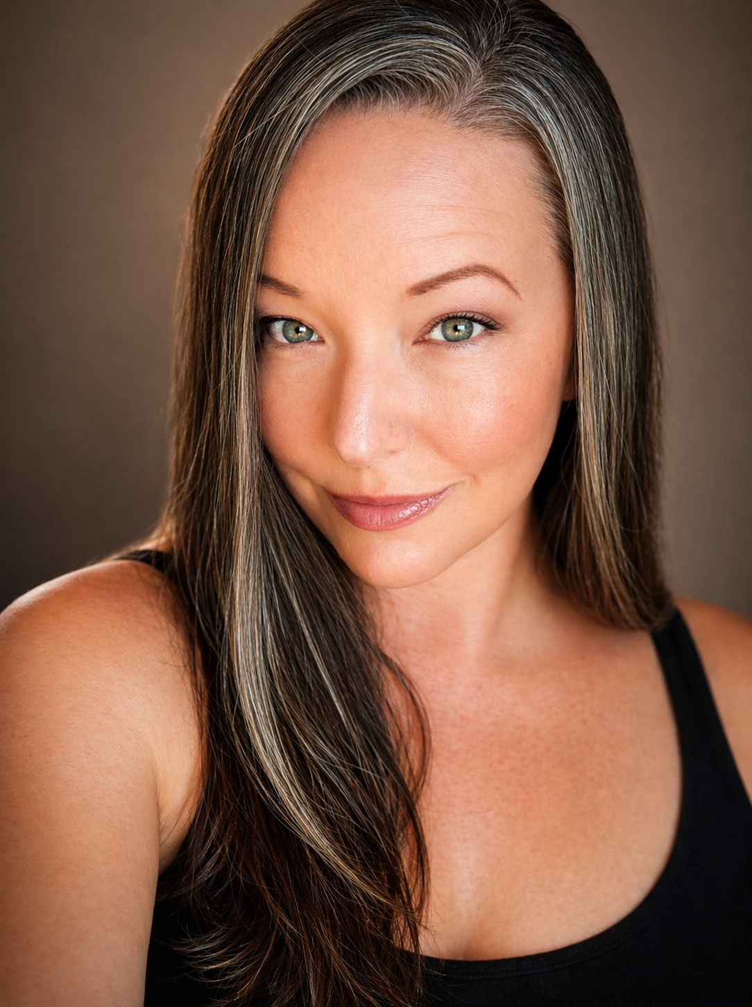 A woman with long brown hair and light makeup smiling softly, wearing a black tank top against a dark background.