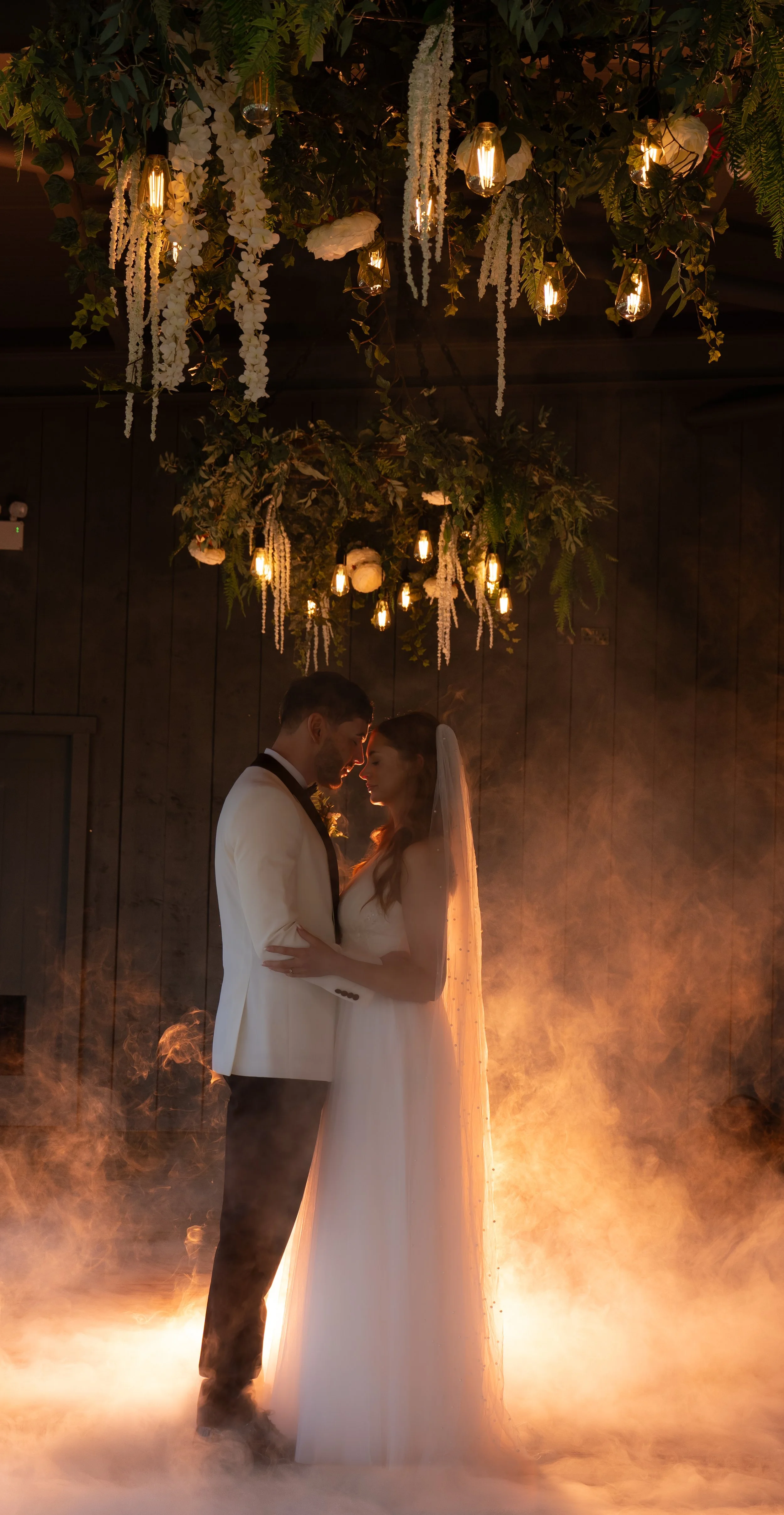 A bride and groom standing close together and touching foreheads under a decorated ceiling with hanging lights and greenery. The scene has warm orange lighting and a foggy atmosphere.