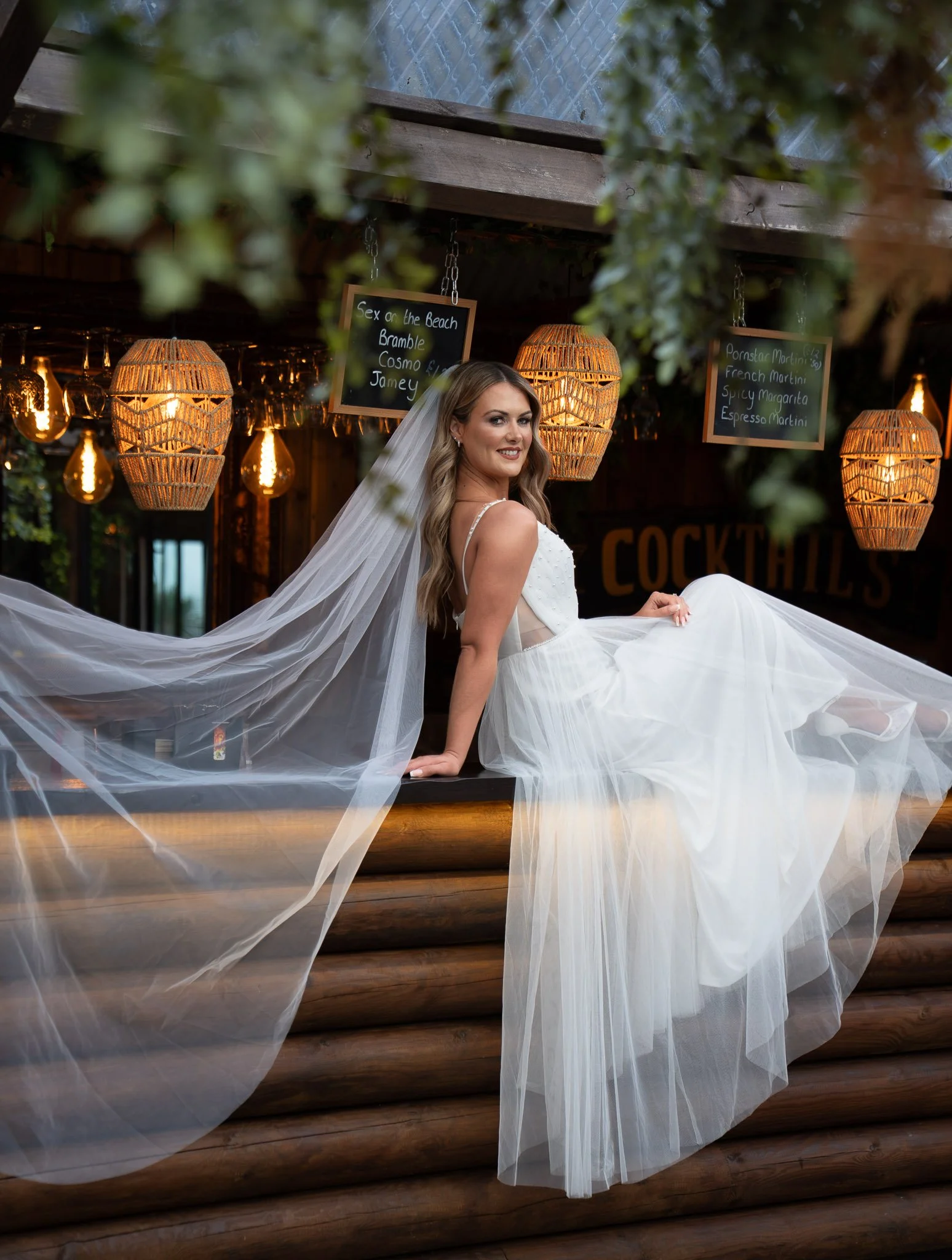 A woman in a white wedding dress with a long veil, sitting on a wooden bar counter with a drink in hand, smiling at the camera, illuminated by warm hanging lights in a bar or restaurant setting.