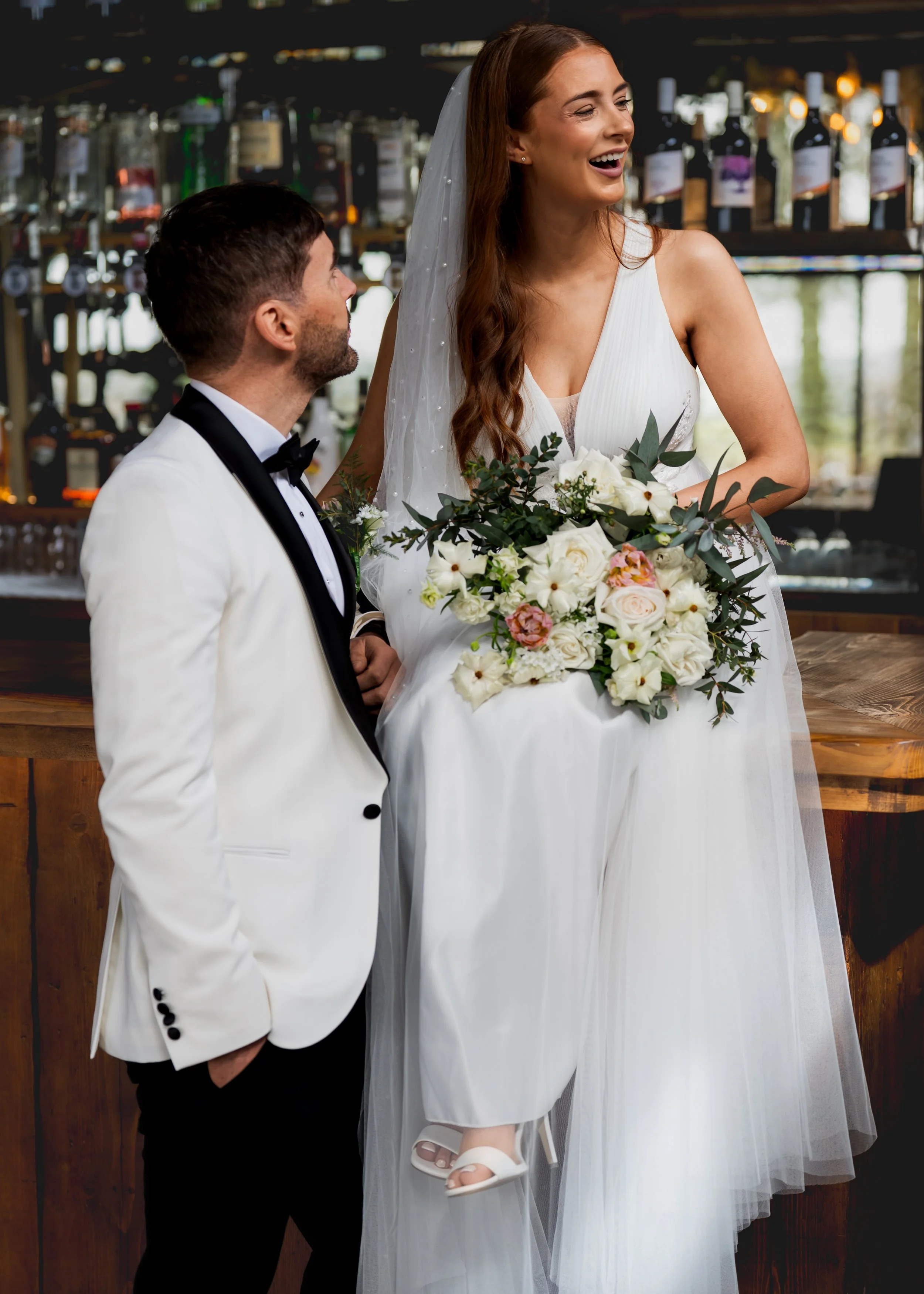 A bride in a white wedding dress holding a bouquet of white and pink flowers, smiling, and a groom in a white tuxedo with a black bow tie, looking at her, in a bar setting with liquor bottles in the background.