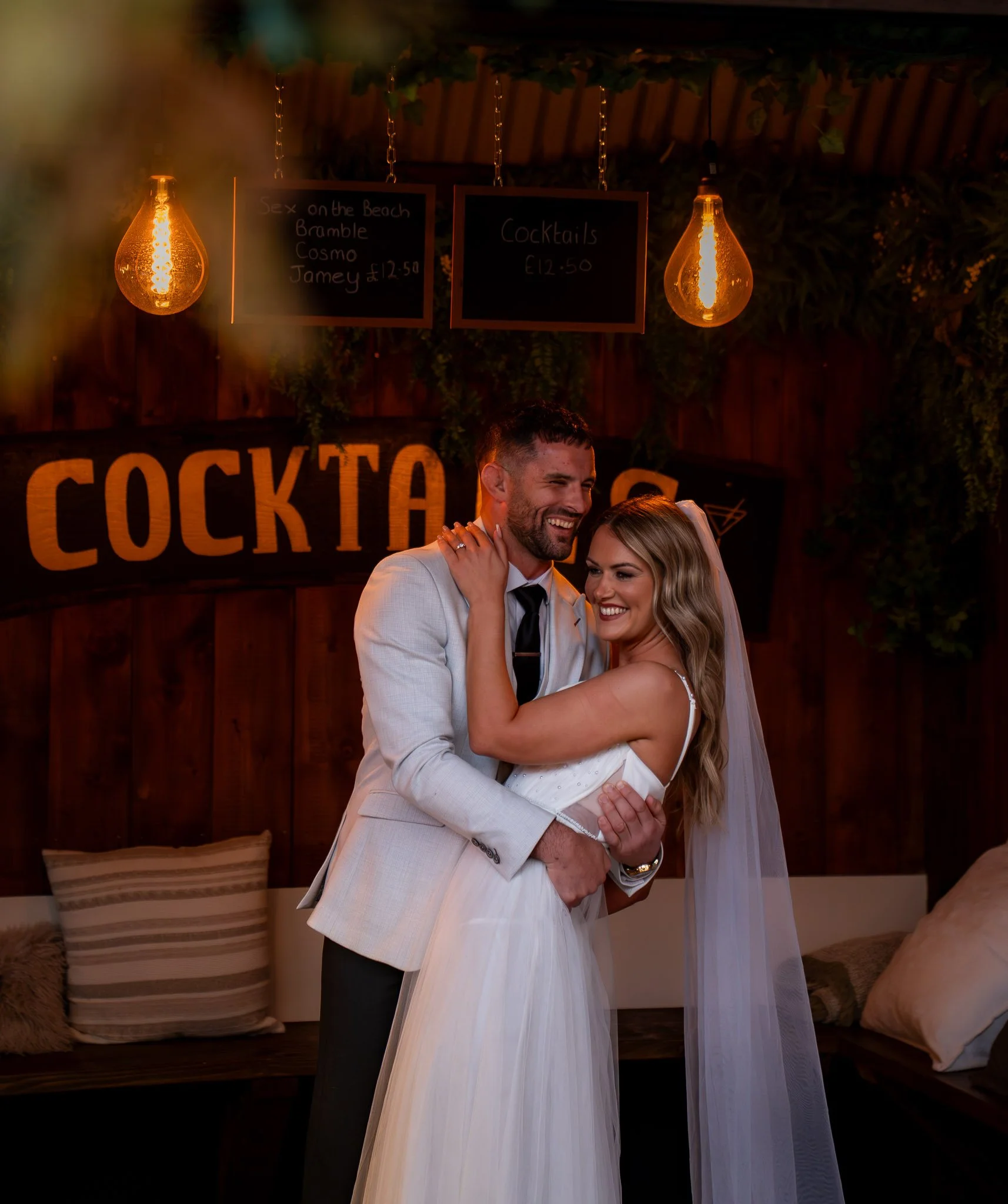 A smiling bride and groom dancing at their wedding reception, with dark wood walls and warm hanging lights in the background.