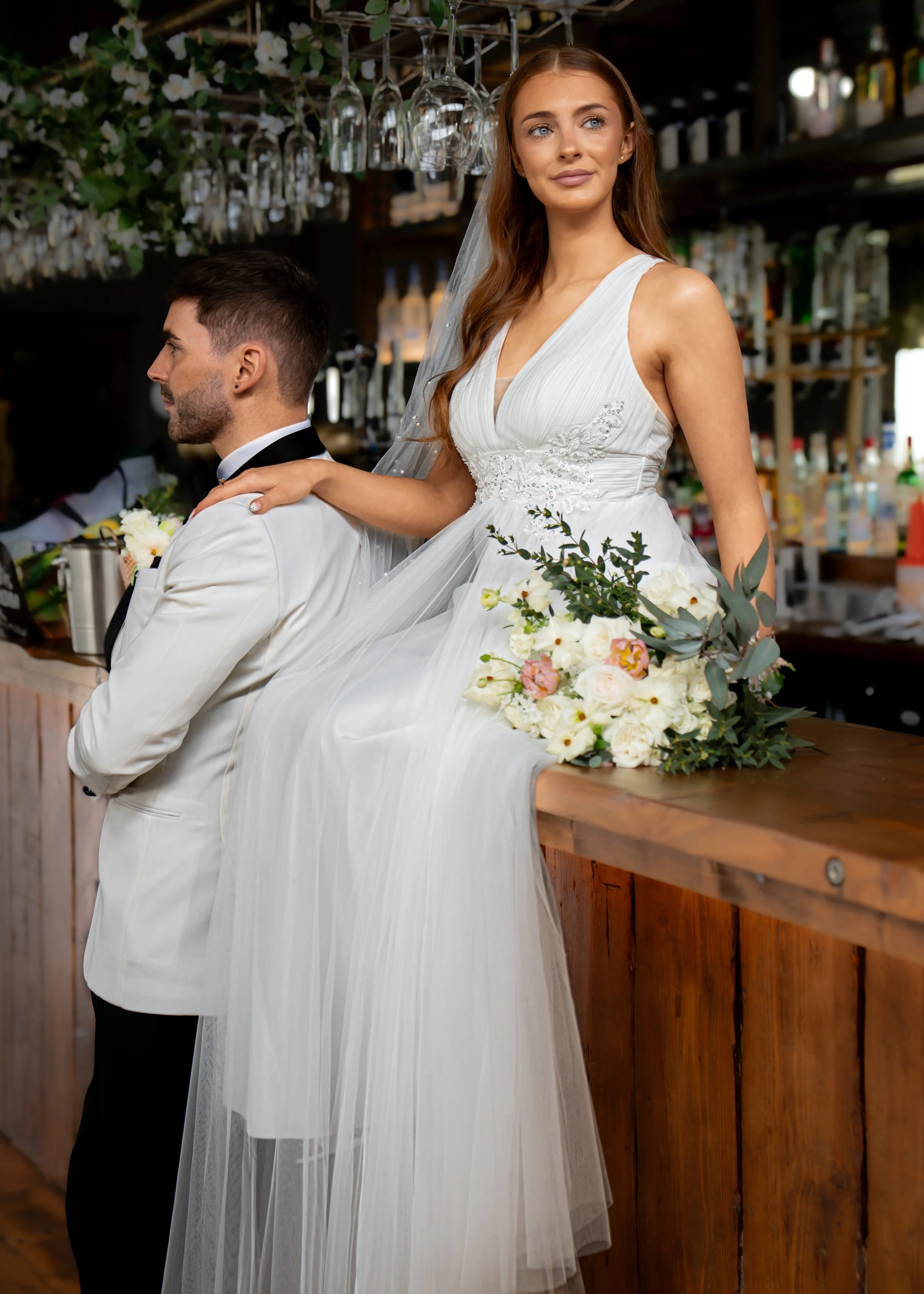 A bride in a white wedding dress with intricate embroidery, standing behind a groom in a white tuxedo with a black bow tie, in a rustic bar setting with hanging glassware and bottles in the background. The bride has long, wavy auburn hair and is hold
