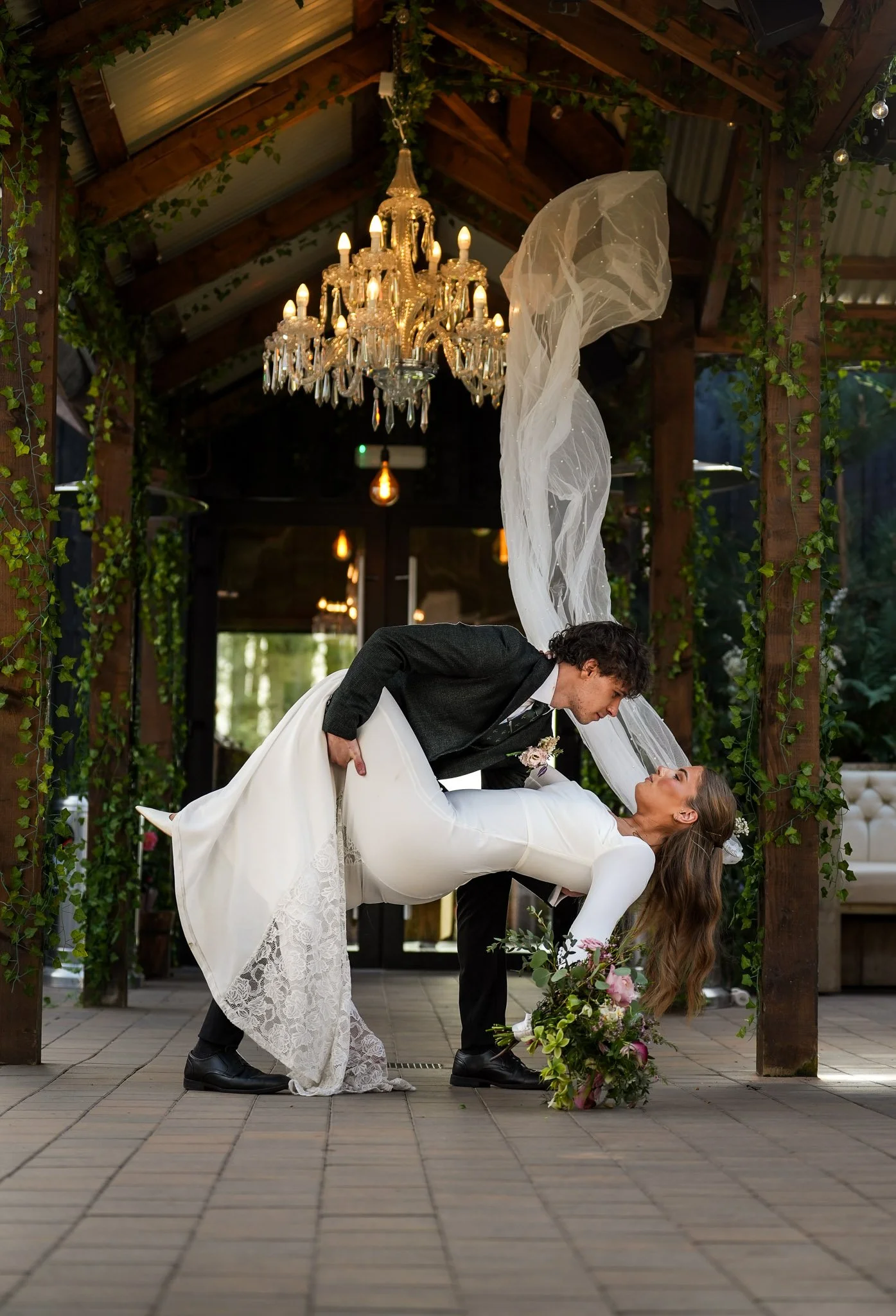 A bride and groom dancing beneath a chandelier, with the groom bending the bride backward, holding her hand, and she holds a flower bouquet with pink and white flowers in front of a floral arrangement on the floor, surrounded by greenery and wooden beams.