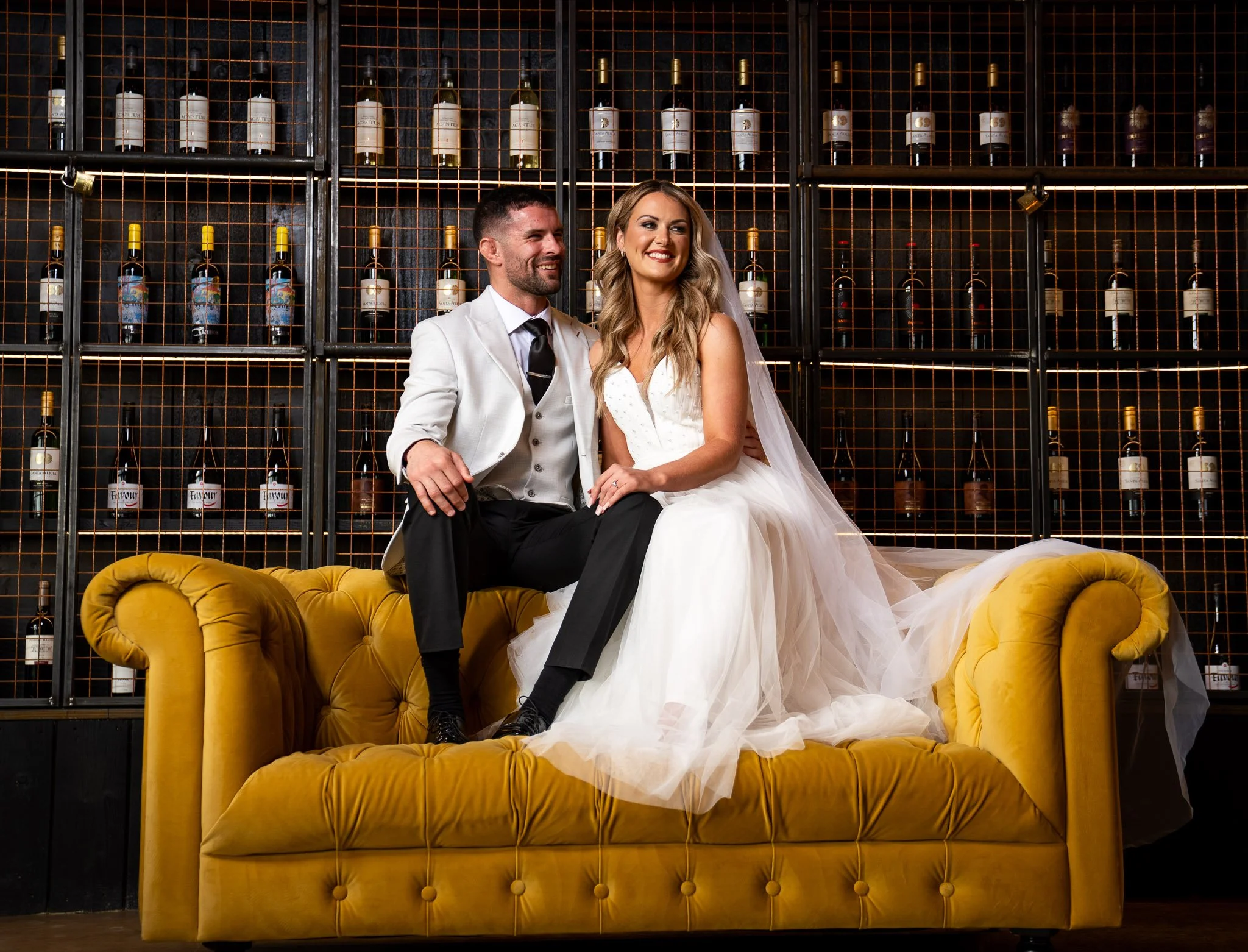 A smiling bride and groom sitting on a yellow velvet sofa in front of a black wall with a metal wine rack filled with bottles of wine.