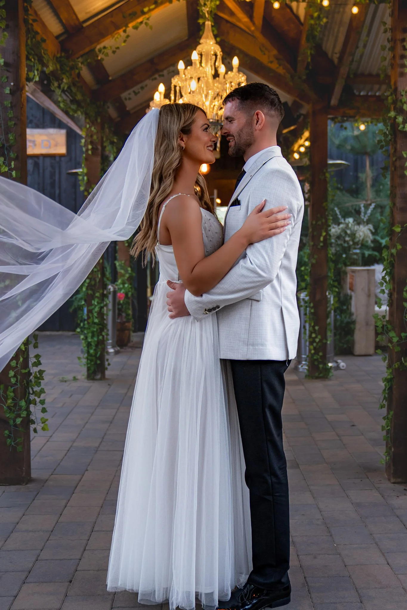 A bride and groom share a moment during their wedding reception under a chandelier in a rustic outdoor setting decorated with greenery.