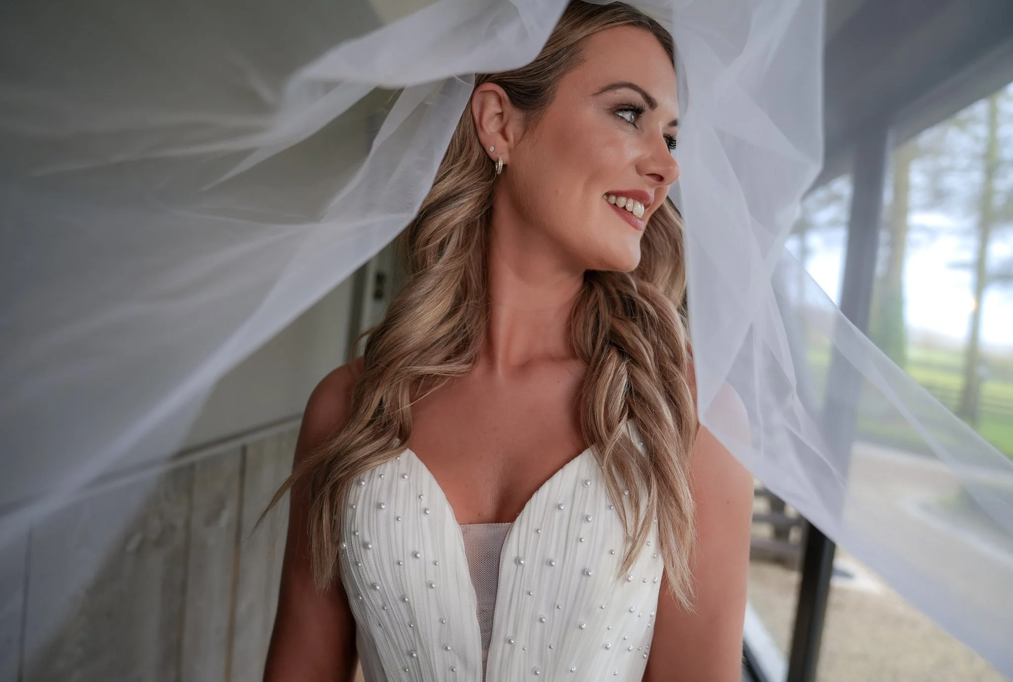 A woman with long wavy hair wearing a white dress with pearl embellishments, smiling and looking to the side, with a tulle veil around her head.