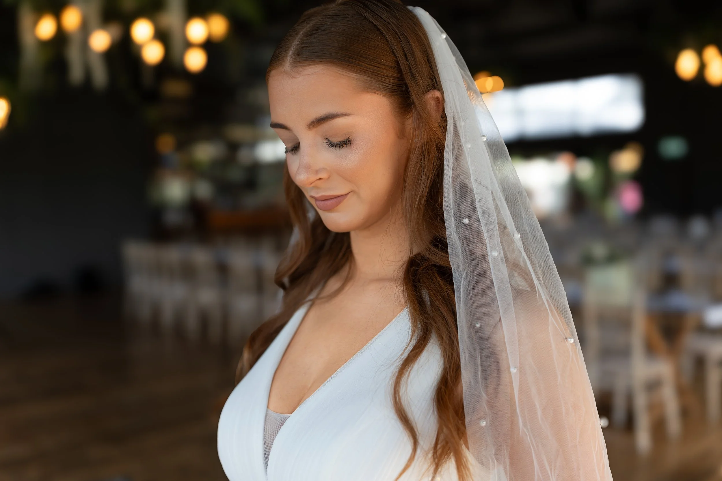 A woman with long brown hair wearing a white dress and a sheer white veil with pearl accents, smiling with her eyes closed in a softly lit indoor setting, possibly a wedding venue.