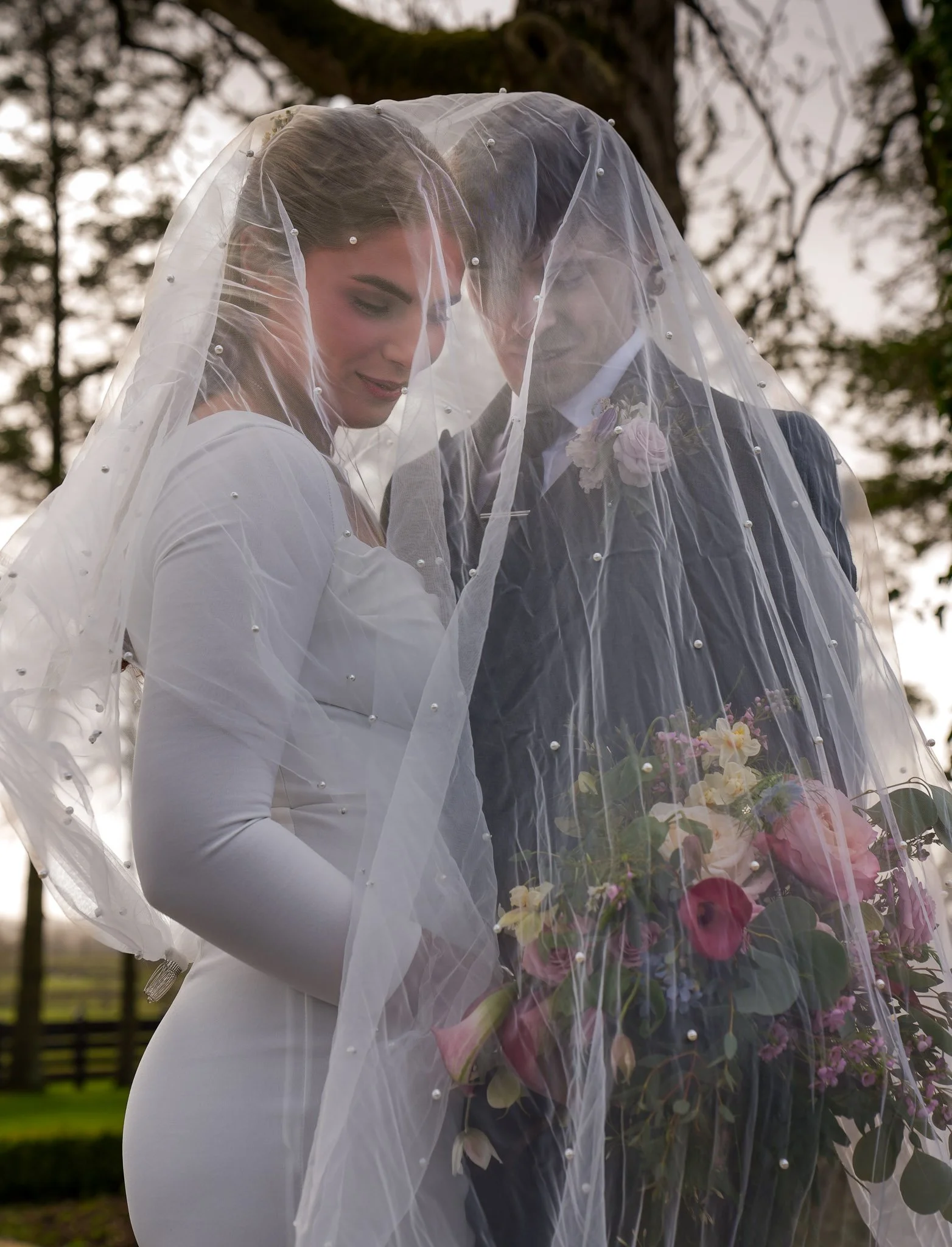 Bride and groom standing together outdoors during daytime, with a wedding veil covering both of their faces, holding a bouquet of flowers.