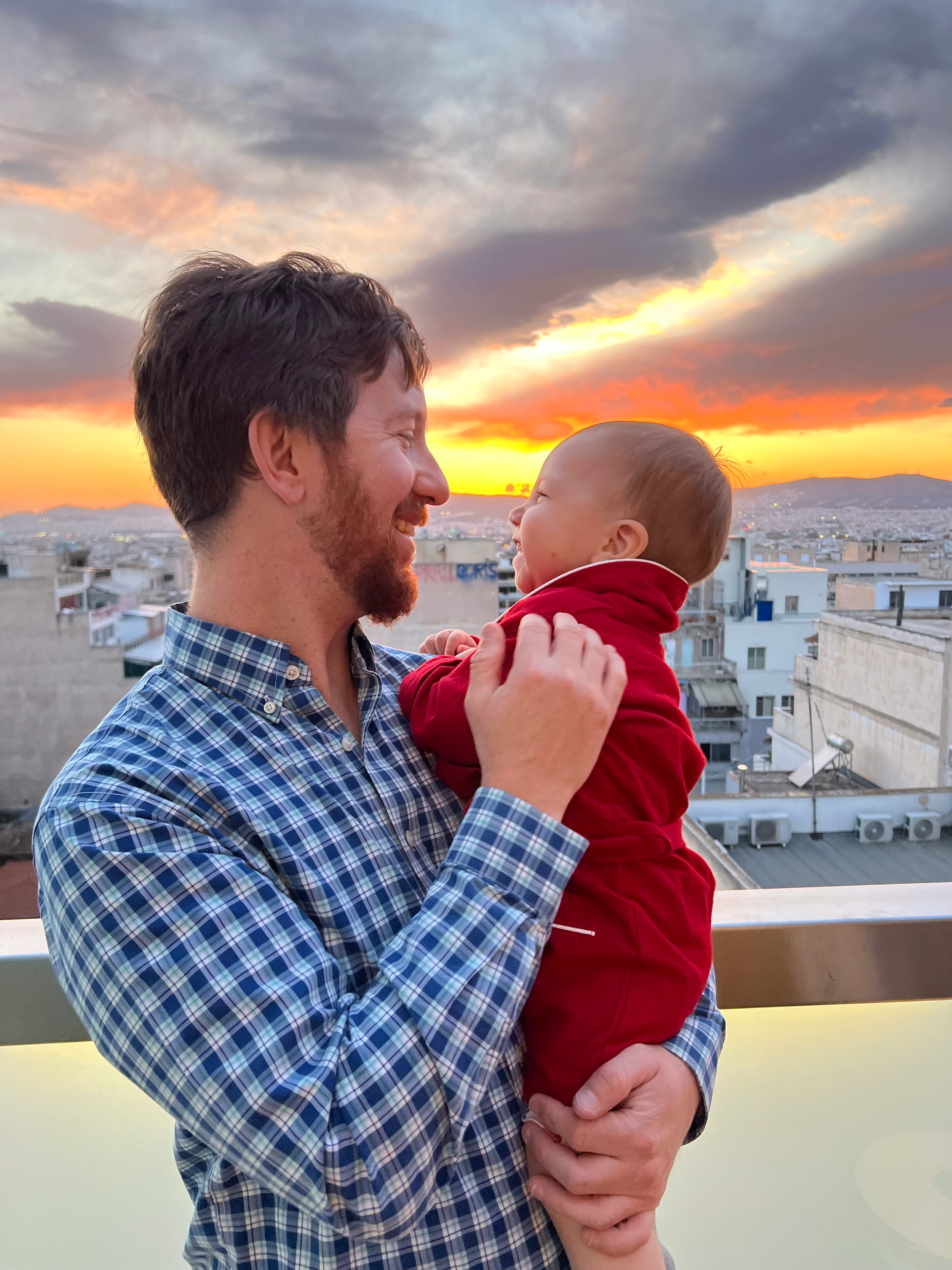 Man holding a smiling baby on a rooftop during a sunset with clouds and city skyline in the background.
