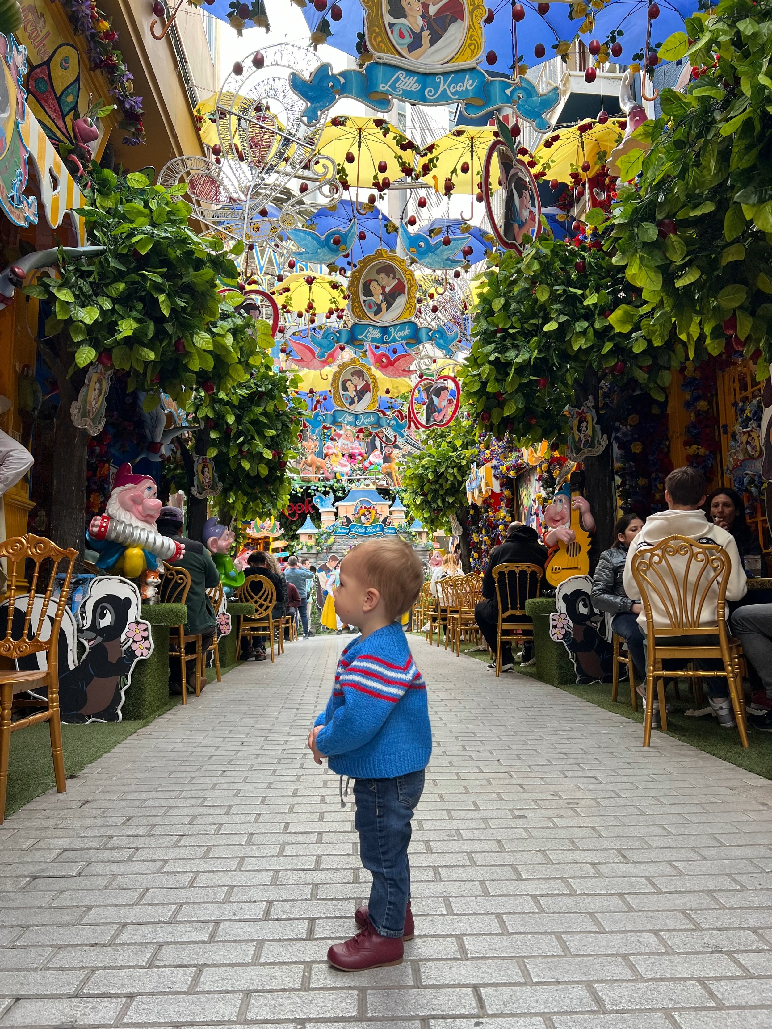 A young boy with a blue sweater and brown boots standing in the middle of a colorful street decorated with cartoon characters, trees with red apples, and bright yellow and blue umbrellas. There are people sitting on golden chairs on either side of the street, and various decorative hanging items overhead, including images of Disney characters and the name "Little Kook."