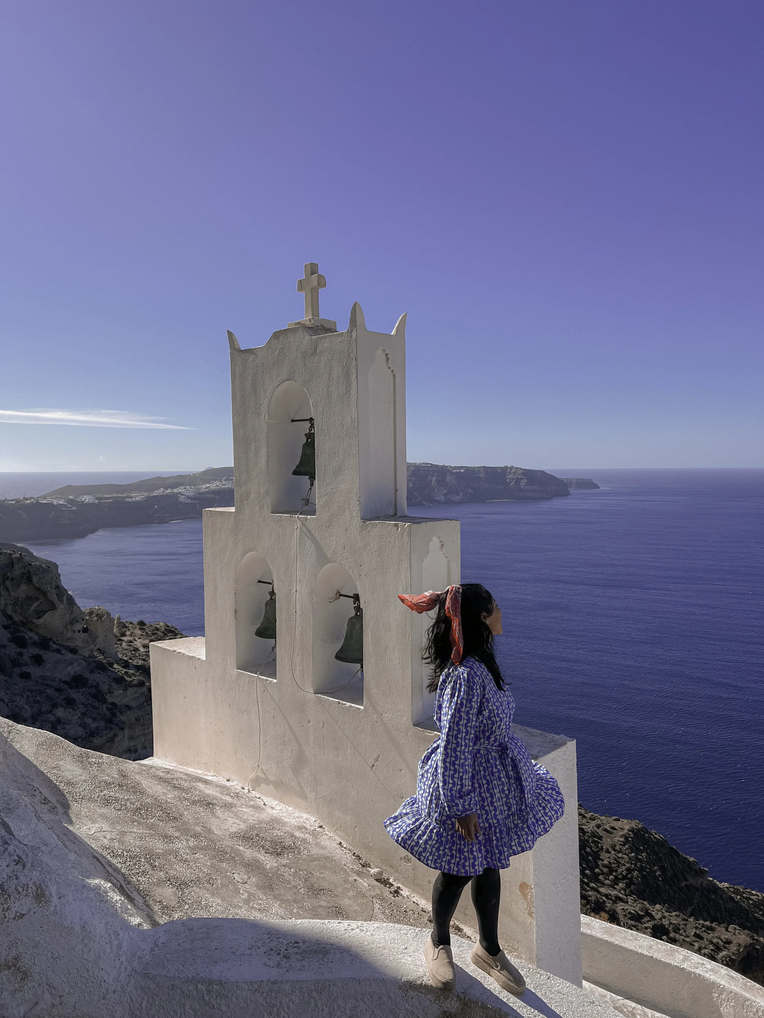 A woman wearing a blue patterned dress, a red scarf, and black tights walking along a white pathway with a white church tower featuring a cross and three bells in front of a blue ocean and sky coastline.