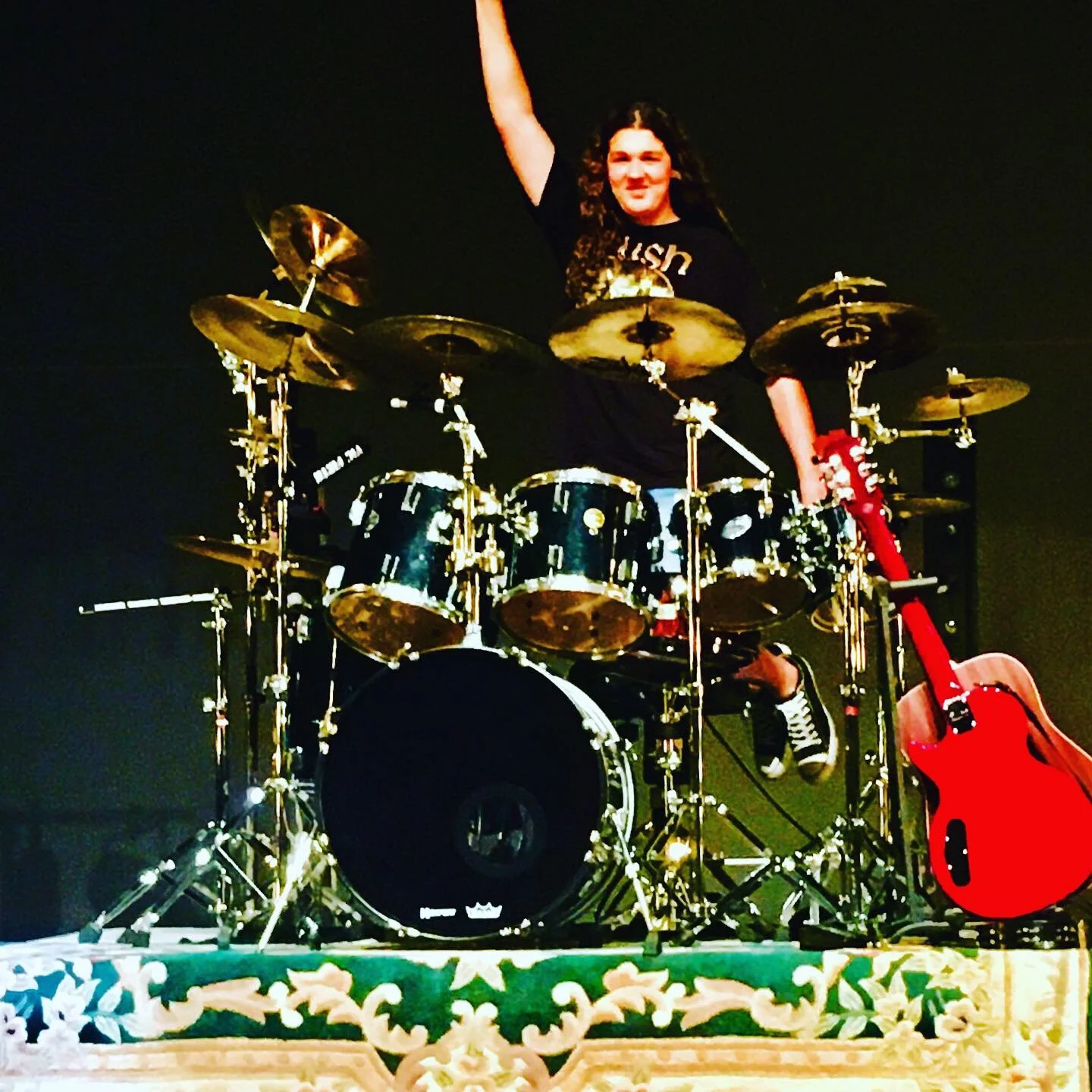 A drummer with long curly hair sitting at a drum set with a guitar leaning against it. He is smiling and raising his right arm, wearing a black RUSH t-shirt, and black sneakers with white stripes.