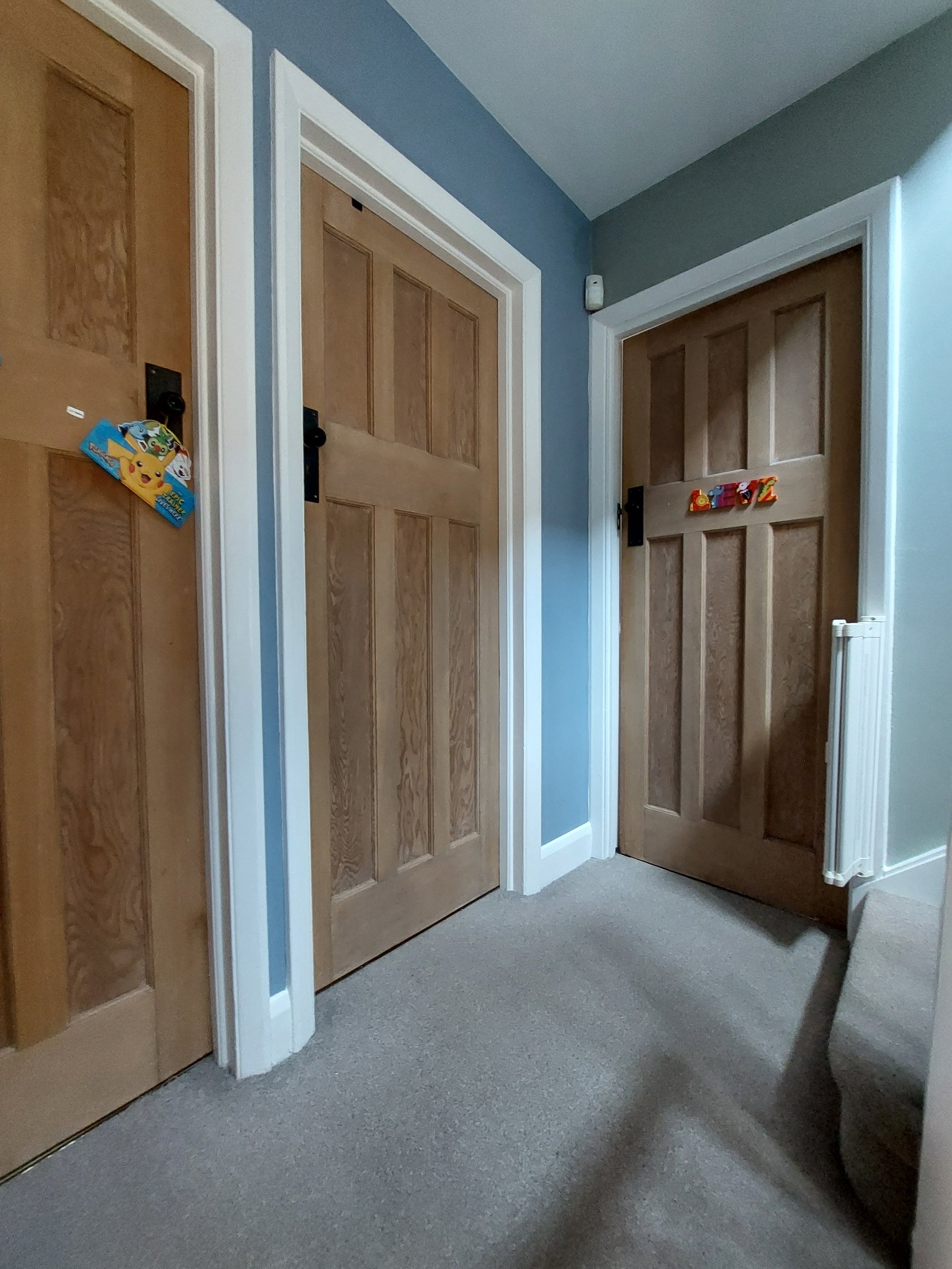A hallway with three wooden doors, two closed and one partially open. The doors have black handles and are framed with white trim. The walls are painted in a two-tone color scheme, with blue on one wall and a darker color on the corner wall. The floor is carpeted in gray, and there is a small white radiator on the right side of the image.