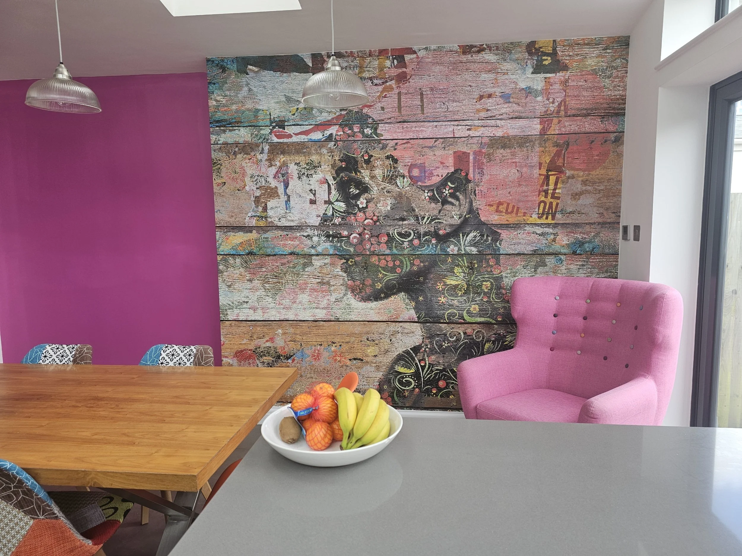 Interior of a dining area with a wooden table, a bowl of fruit, pink armchair, featuring a colourful wallpapered art mural, pink painted wall, hanging light fixtures, and a doorway with window.
