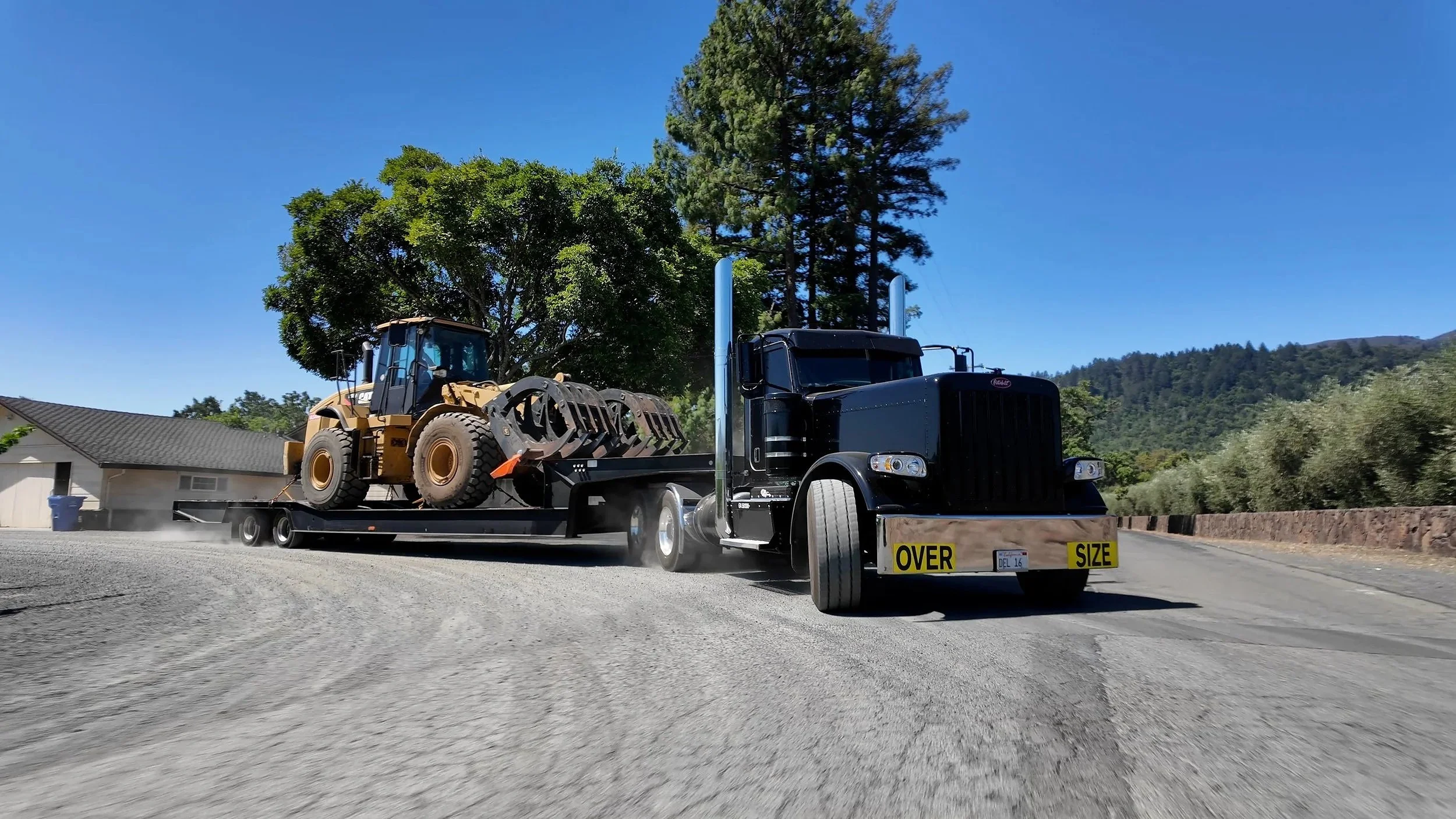 A black semi-truck hauling a construction vehicle on a flatbed trailer labeled 'OVER SIZE'. Heavy Equipment hauling Experts Northern California