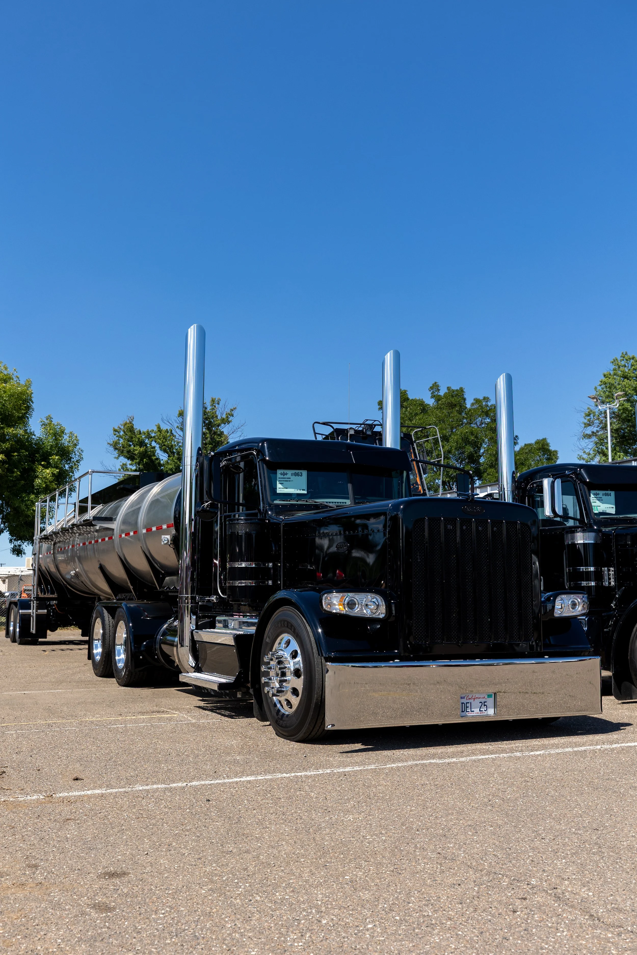 Black semi-truck with a silver tanker trailer parked in an outdoor lot, with green trees and a clear blue sky in the background. Wine transportation Services Napa Valley
