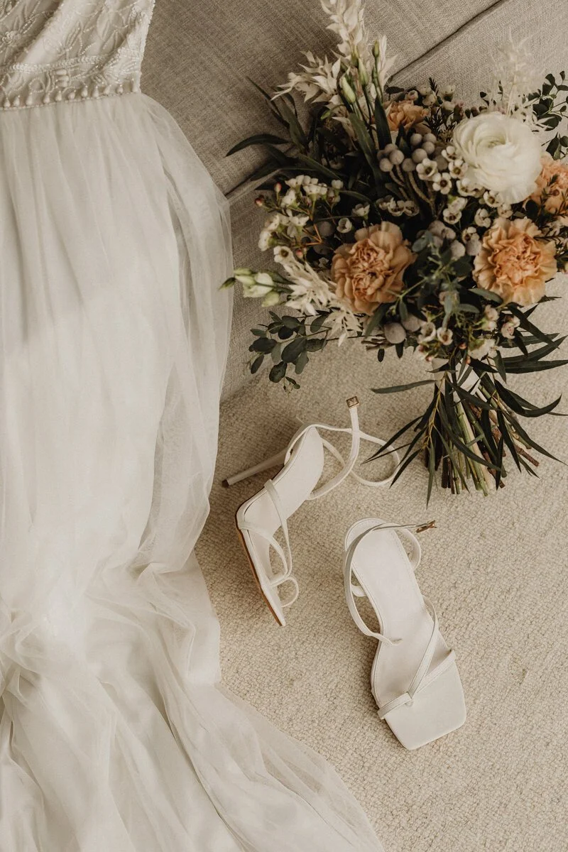 A wedding dress, a pair of white high-heeled shoes, and a bouquet of flowers on a beige carpeted floor.