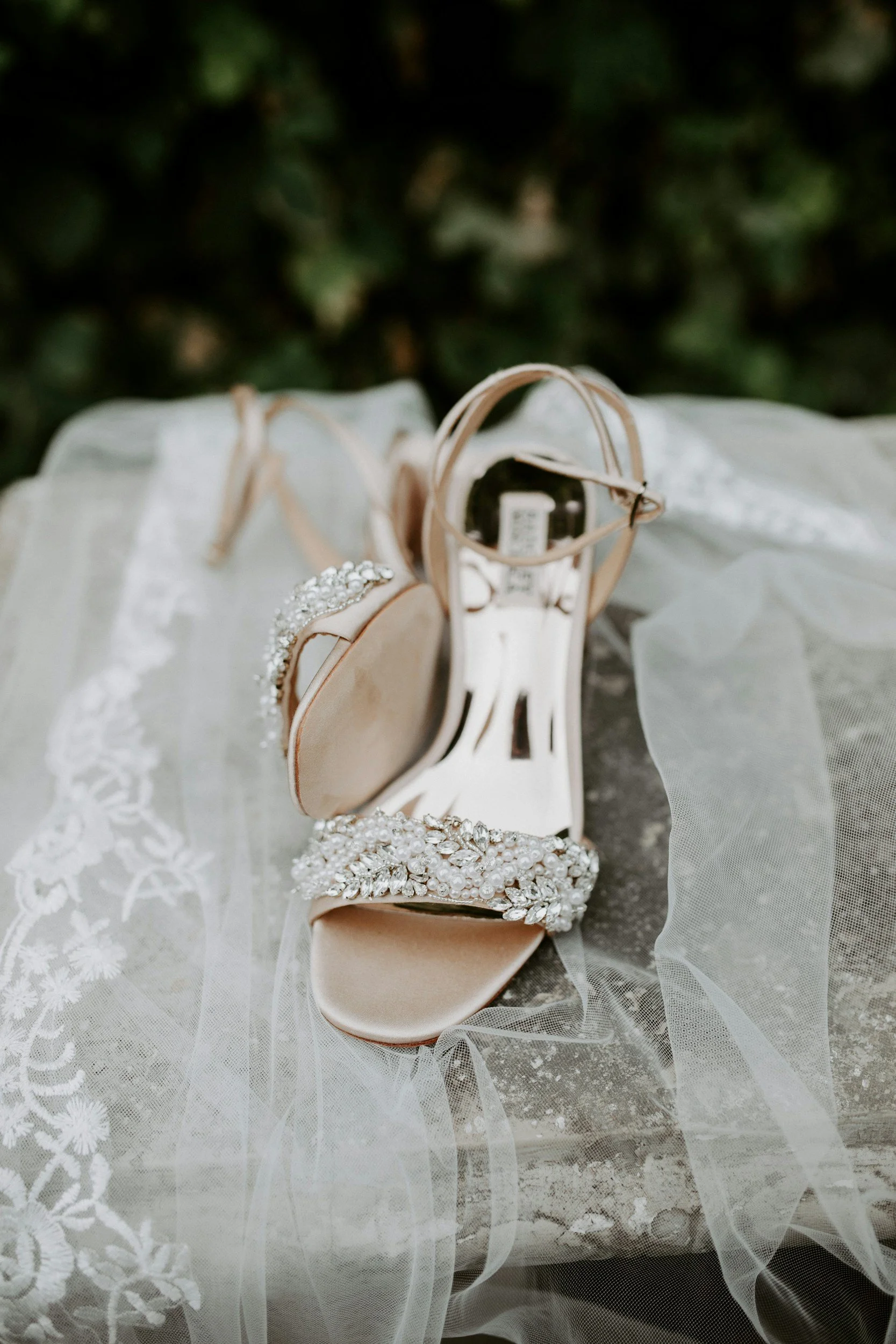 A pair of beige high heel shoes with ankle straps, embellished with rhinestones, resting on a lace fabric and polished gray surface.