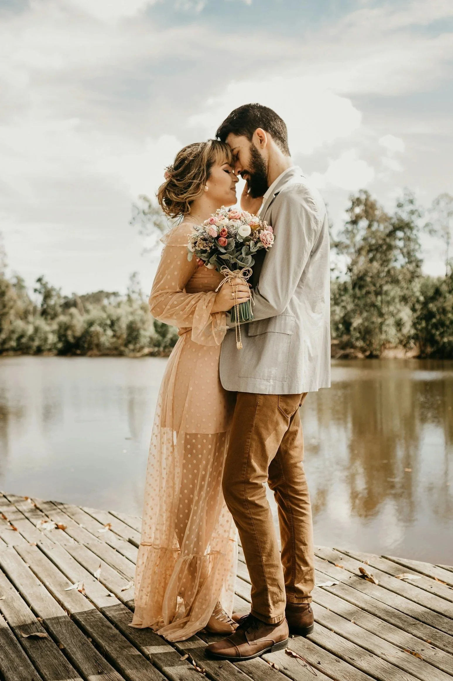 A couple stands close to each other on a wooden dock by a body of water, with their foreheads touching and eyes closed, holding a bouquet of flowers. The woman is wearing a light peach, dotted, sheer dress, and the man is dressed in a light-colored blazer with brown pants.