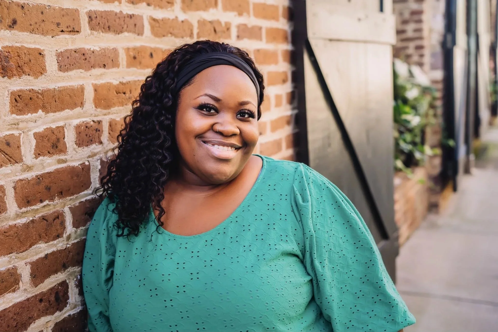 A woman with curly black hair and a black headband smiling, wearing a teal blouse, sitting against a brick wall outside.