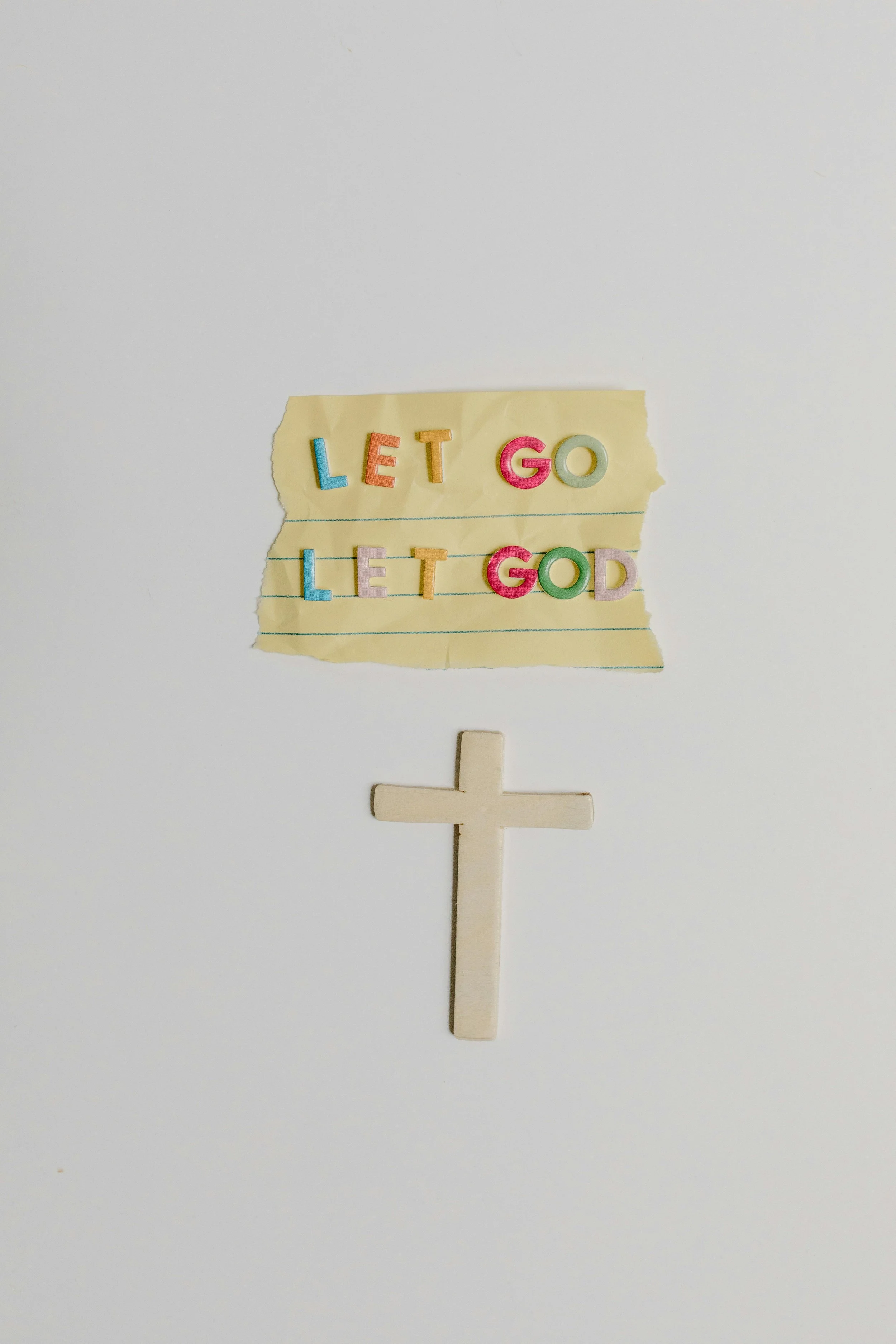A small sign made from colorful letter magnets on lined yellow paper displays the phrase 'Let Go Let God,' with a white cross below it, set against a white background.