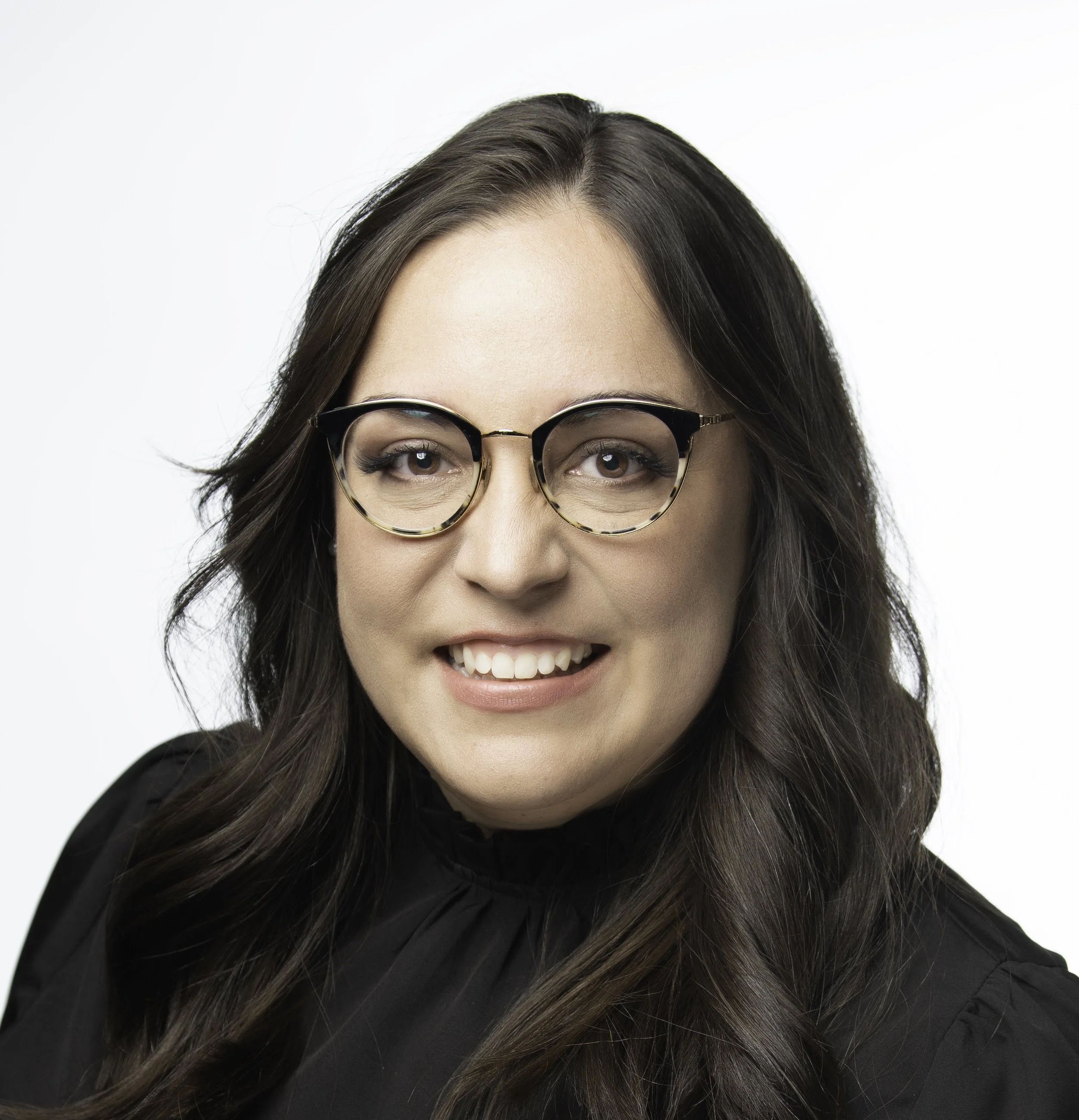 Close-up of Lili-Erika Zuno with brown hair, wearing glasses with tortoise-shell frames, smiling with visible teeth, against a white background.
