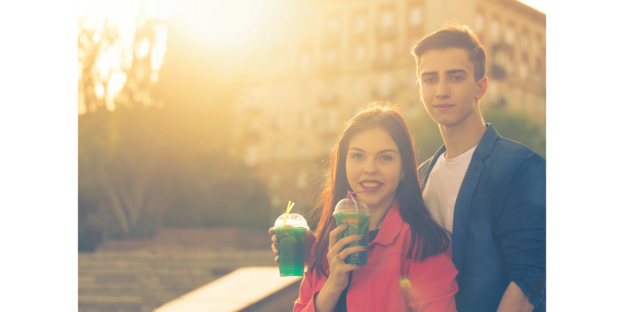 Young man and woman standing outdoors at sunset, holding colorful drinks and smiling at the camera.