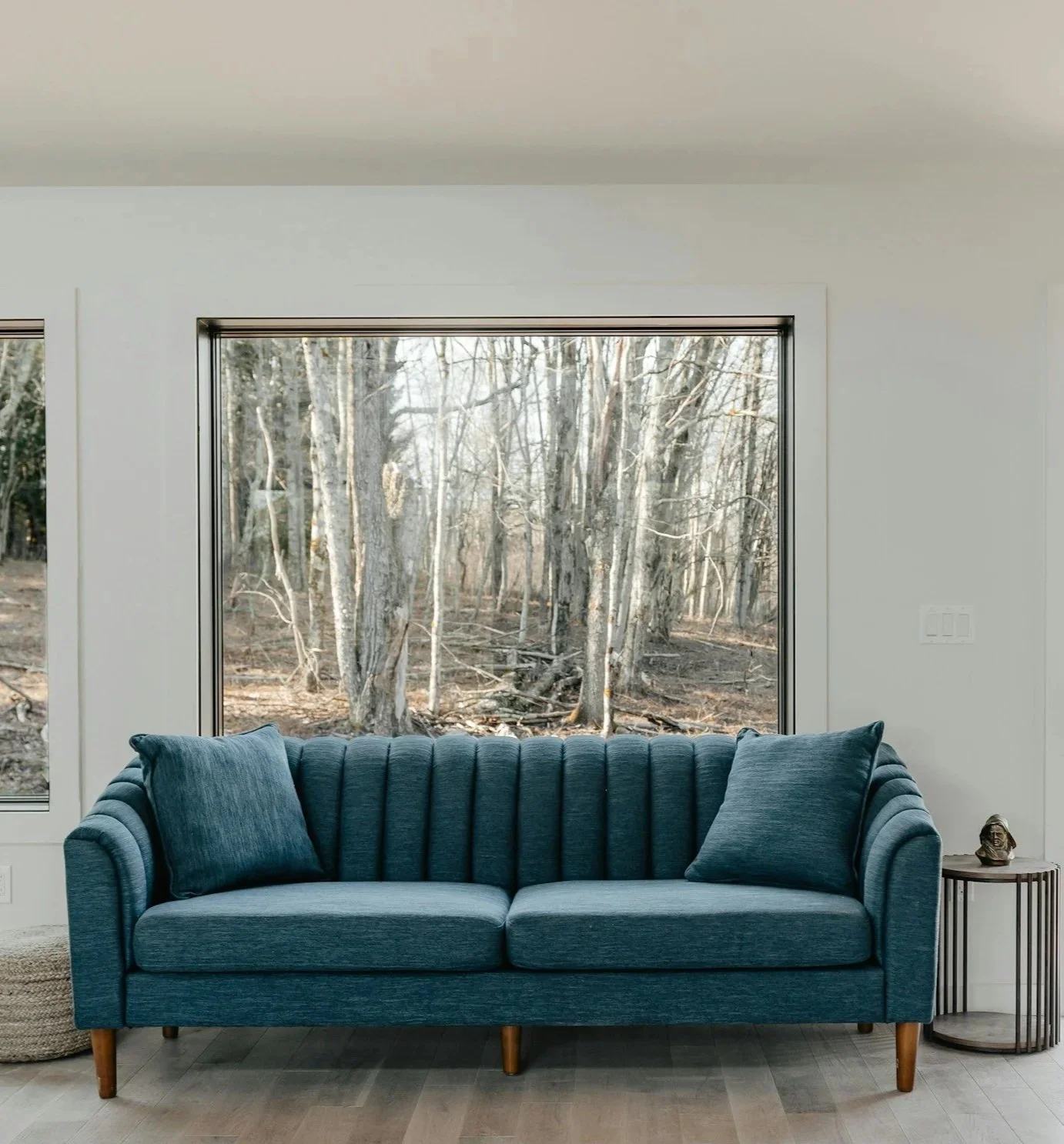 A blue upholstered sofa with pillows inside a living room, in front of large window showing a wooded outdoor scene.
