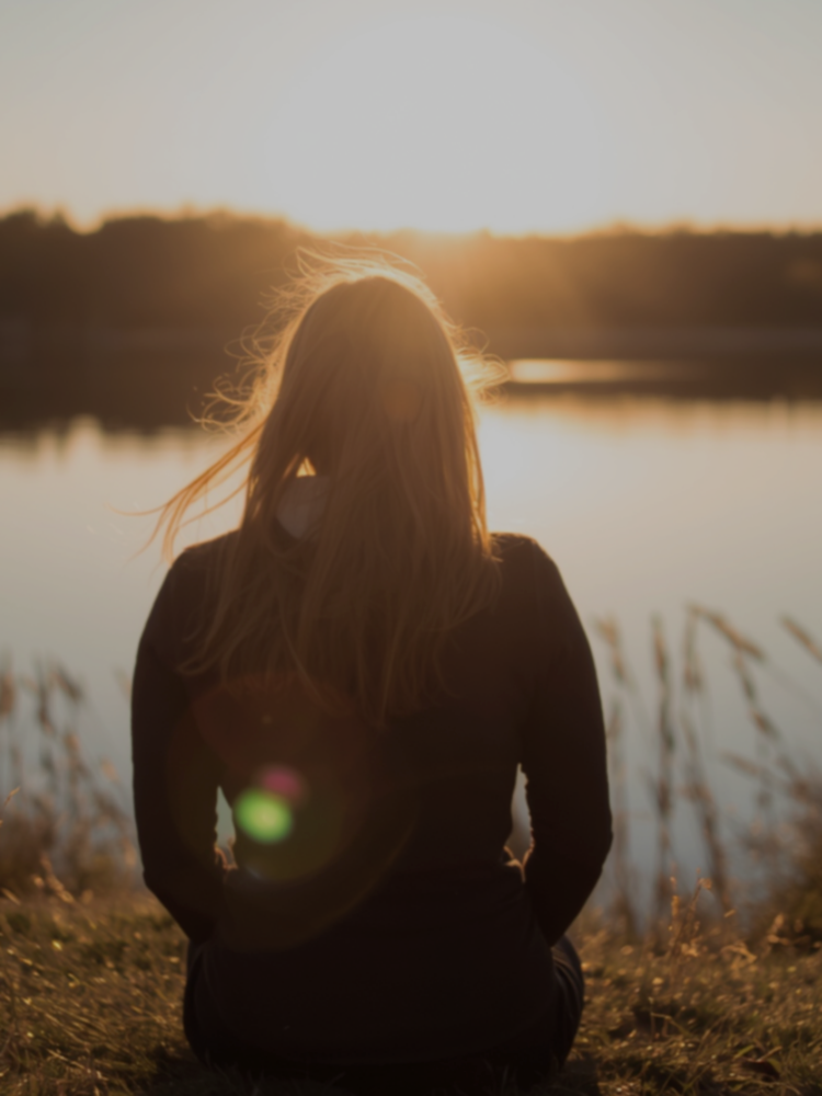 A woman sitting outdoors by a lake at sunset, facing away from the camera.