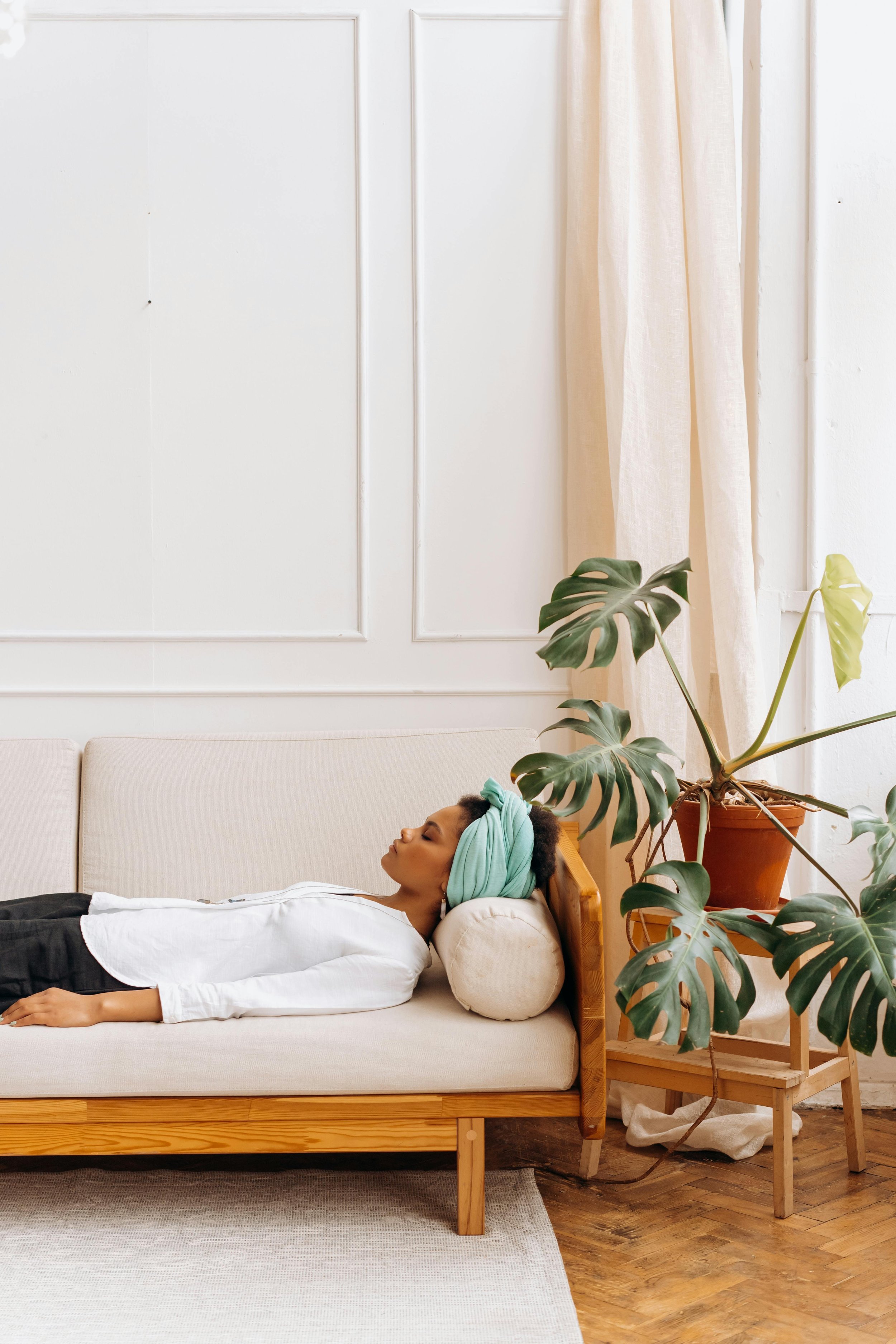 A woman with a towel wrapped around her head is sleeping on a bed in a minimalist room with white walls, beige curtains, a cushion under her head, and a large potted Monstera plant beside the bed.
