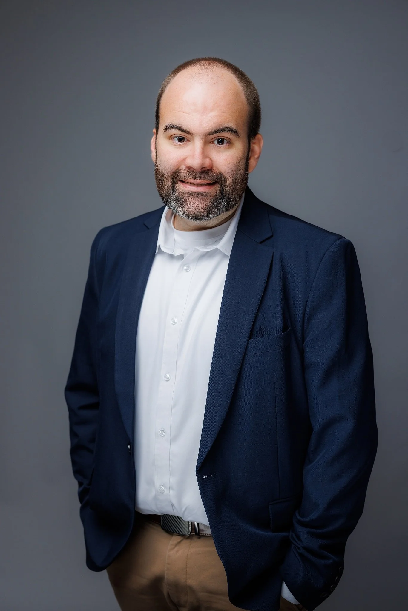 Headshot of a man with a beard, wearing a dark blue blazer, white shirt, and khaki pants, standing against a plain grey background.