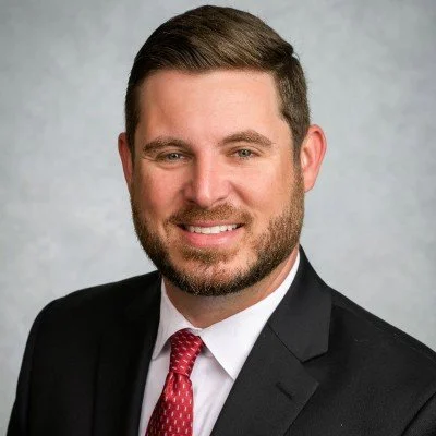 Headshot of a man with short brown hair, beard, wearing a black suit, white shirt, and red patterned tie, smiling against a gray background.