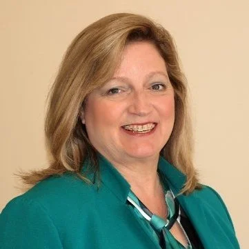 Portrait of a woman with shoulder-length blonde hair, smiling, wearing a teal blazer and patterned blouse against a neutral background.