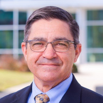 Professional man with glasses wearing a suit and tie, standing outdoors in front of a modern building.