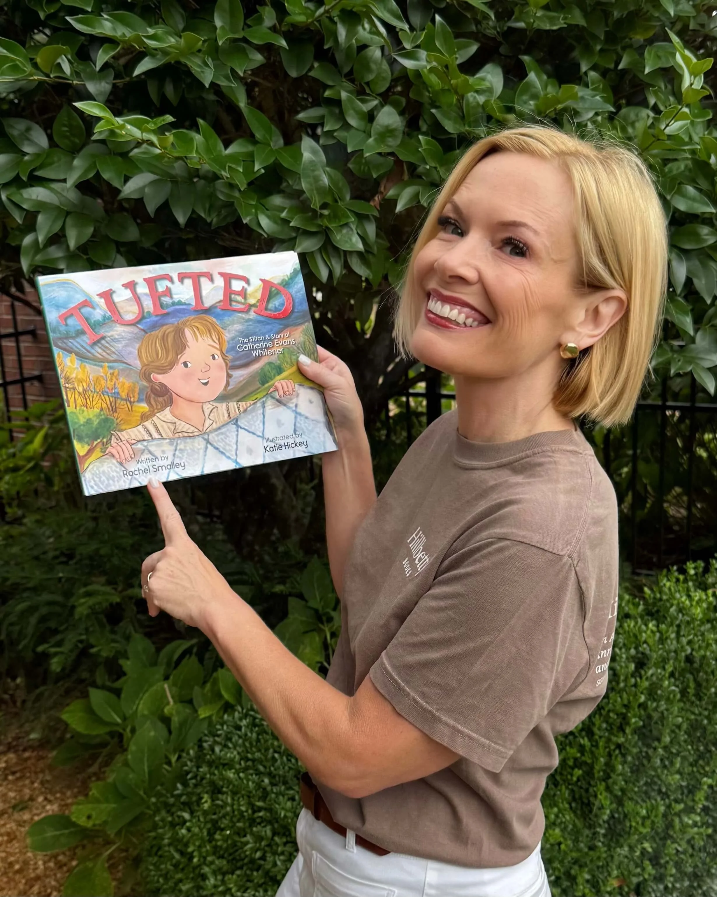 Rachel Smalley smiling with short blonde hair holding a children's book titled "TUFTED" outdoors in front of a leafy green hedge.
