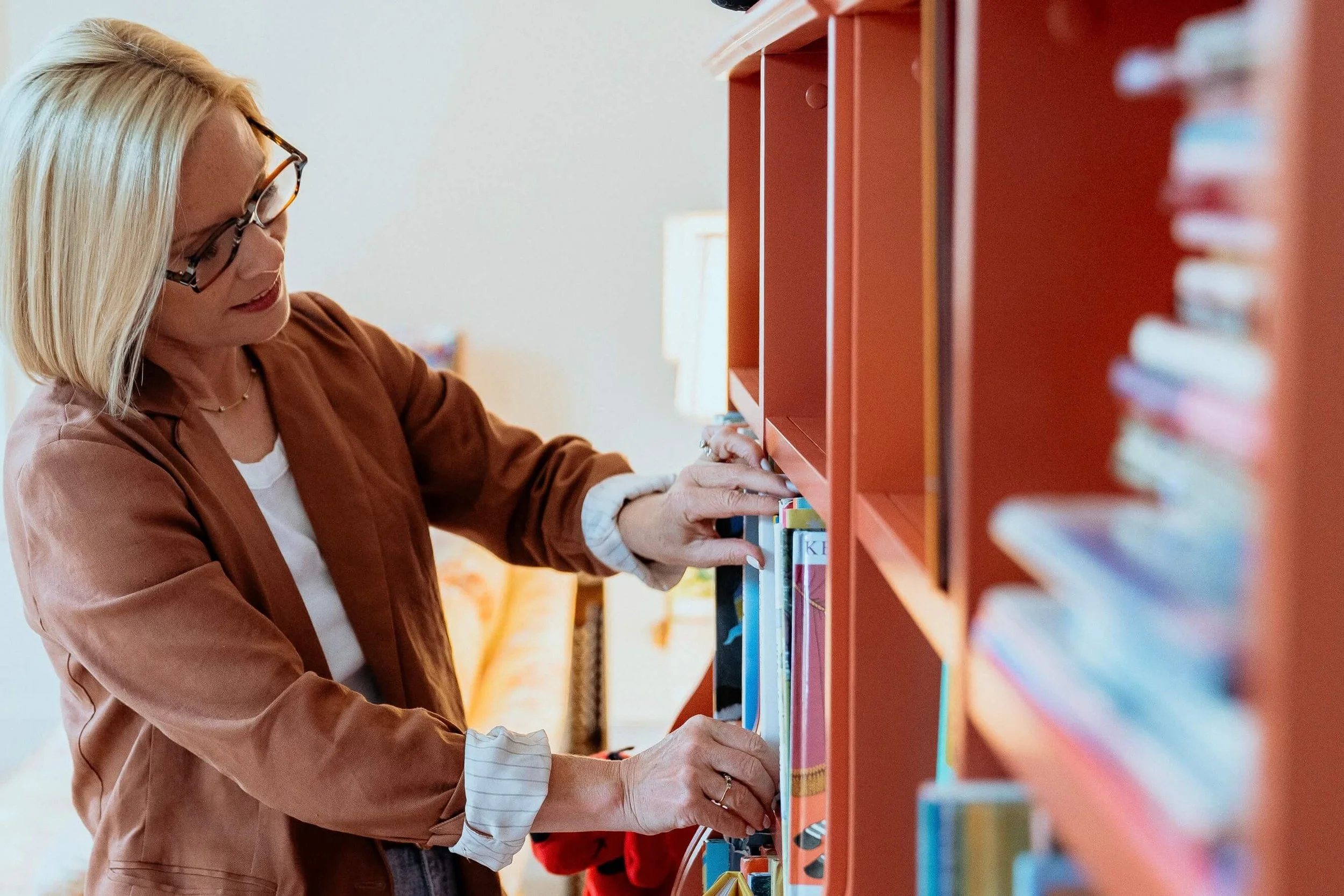 Rachel Smalley with blonde hair and glasses selecting a book from a red bookshelf. She is wearing a brown jacket and a white shirt, and is focused on choosing a book in a well-lit room.
