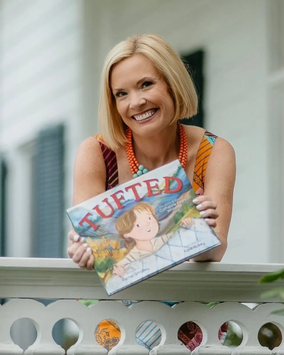 Rachel Smalley smiling and holding a children's book titled 'TUFTED'. She is sitting outside on a porch or balcony with a white railing, wearing a colorful top and a turquoise and orange necklace.