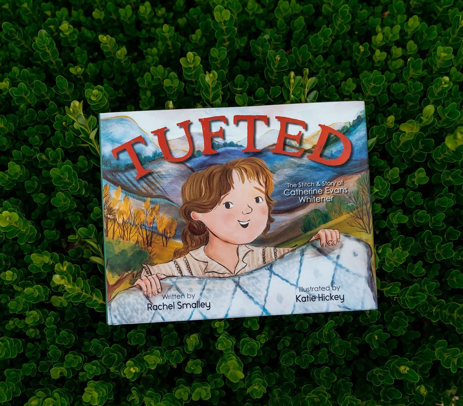 Children's book titled 'TUFTED' resting on green clover leaves, featuring illustrated girl holding a tufted fabric with mountains and trees in the background.