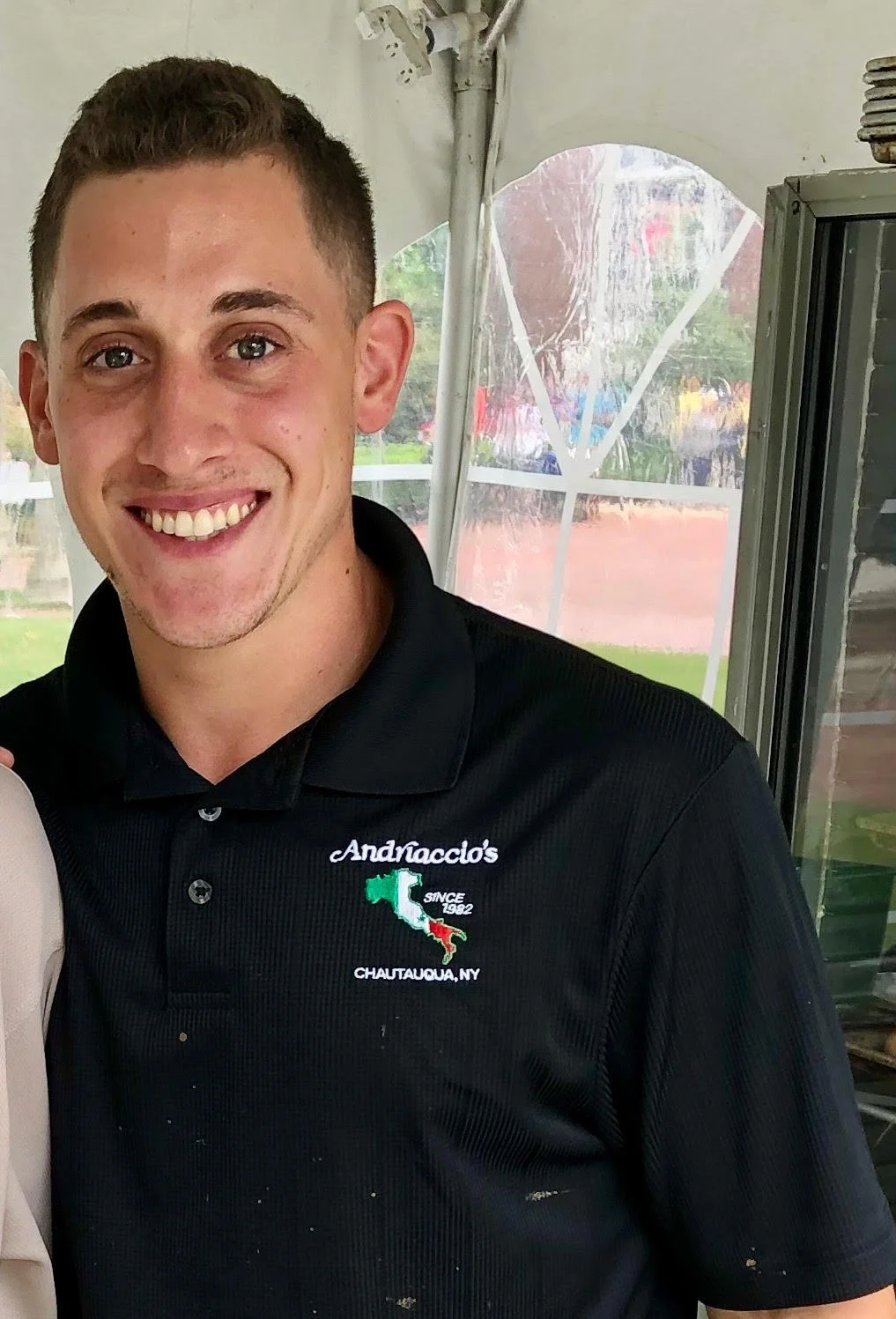 Smiling young man wearing a black polo shirt with embroidery of an Italian flag and the text '&Andraccio's SINCE 1982 CHAUTAUQUA, NY,' indoors with a window and tent behind him.