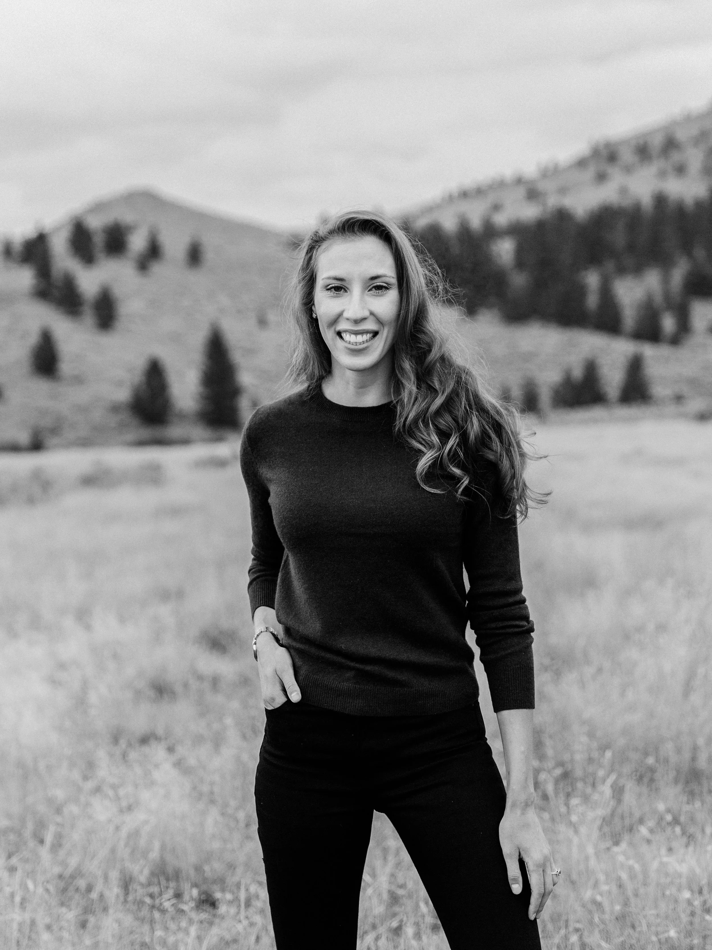 A smiling woman with long wavy hair wearing a dark sweater and pants standing outdoors in a grassy field with hills and trees in the background in black and white.