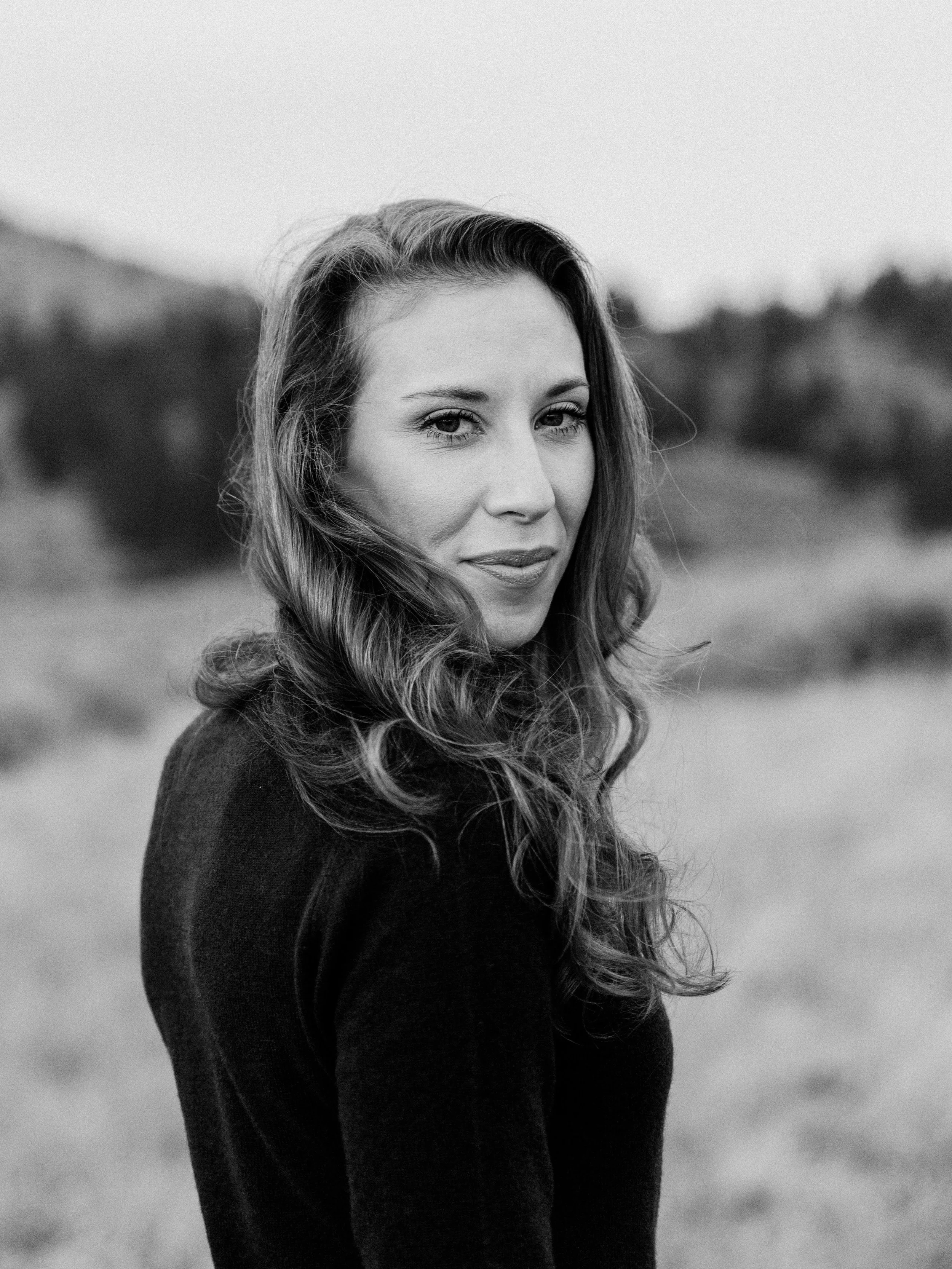Black and white portrait of a woman with long wavy hair, outdoors with blurred natural background.