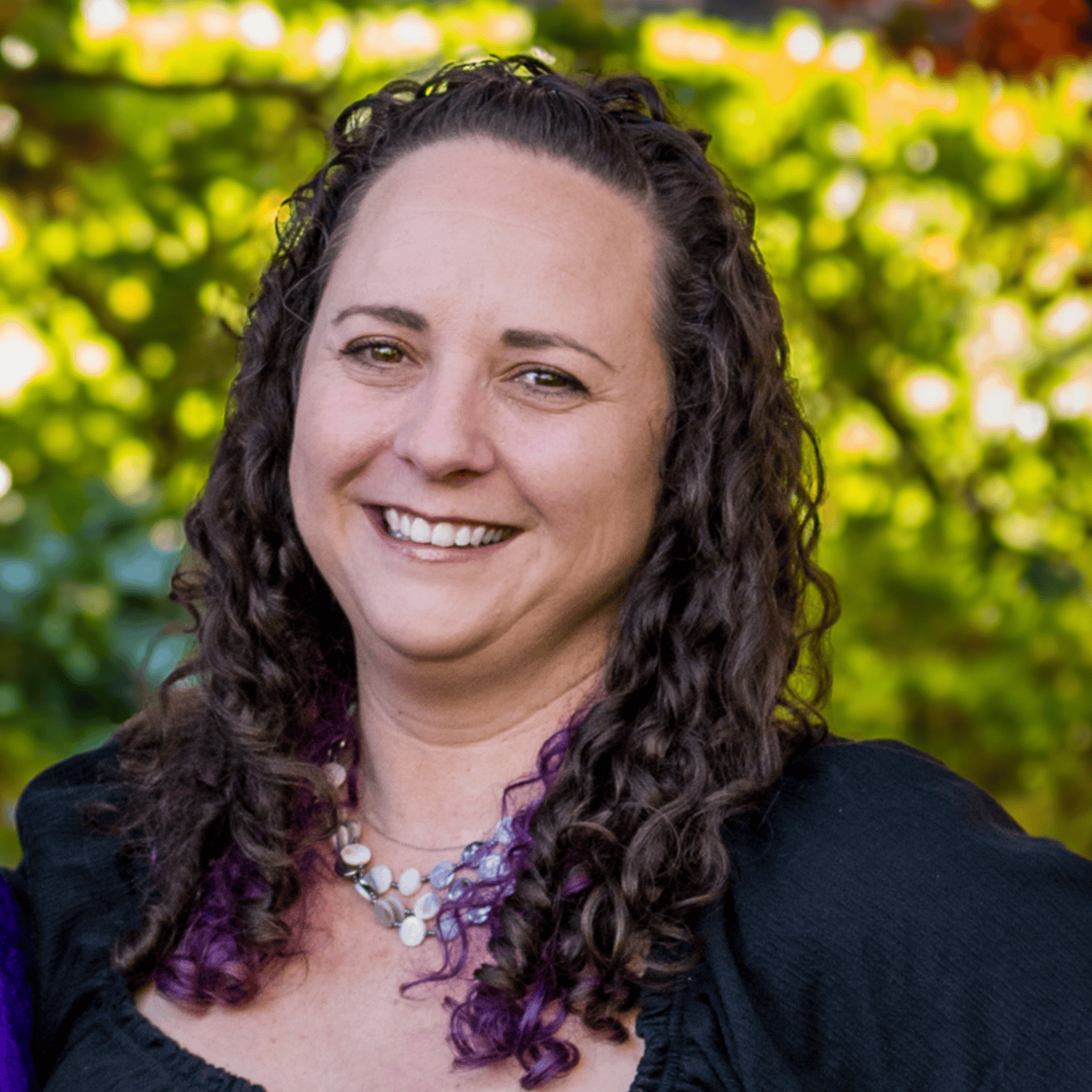 A woman with long curly brown hair smiling outdoors with green blurred trees in the background, wearing a black top, a multi-layerd shell necklace, and purple accents in her hair.