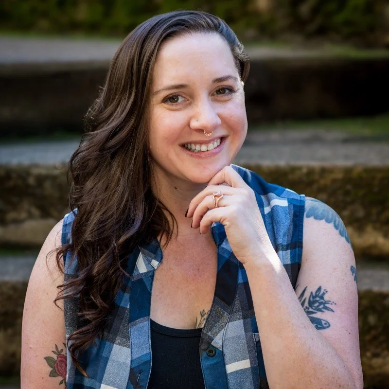 A smiling woman with long dark hair, wearing a blue plaid sleeveless shirt, outdoors with blurred greenery in the background.