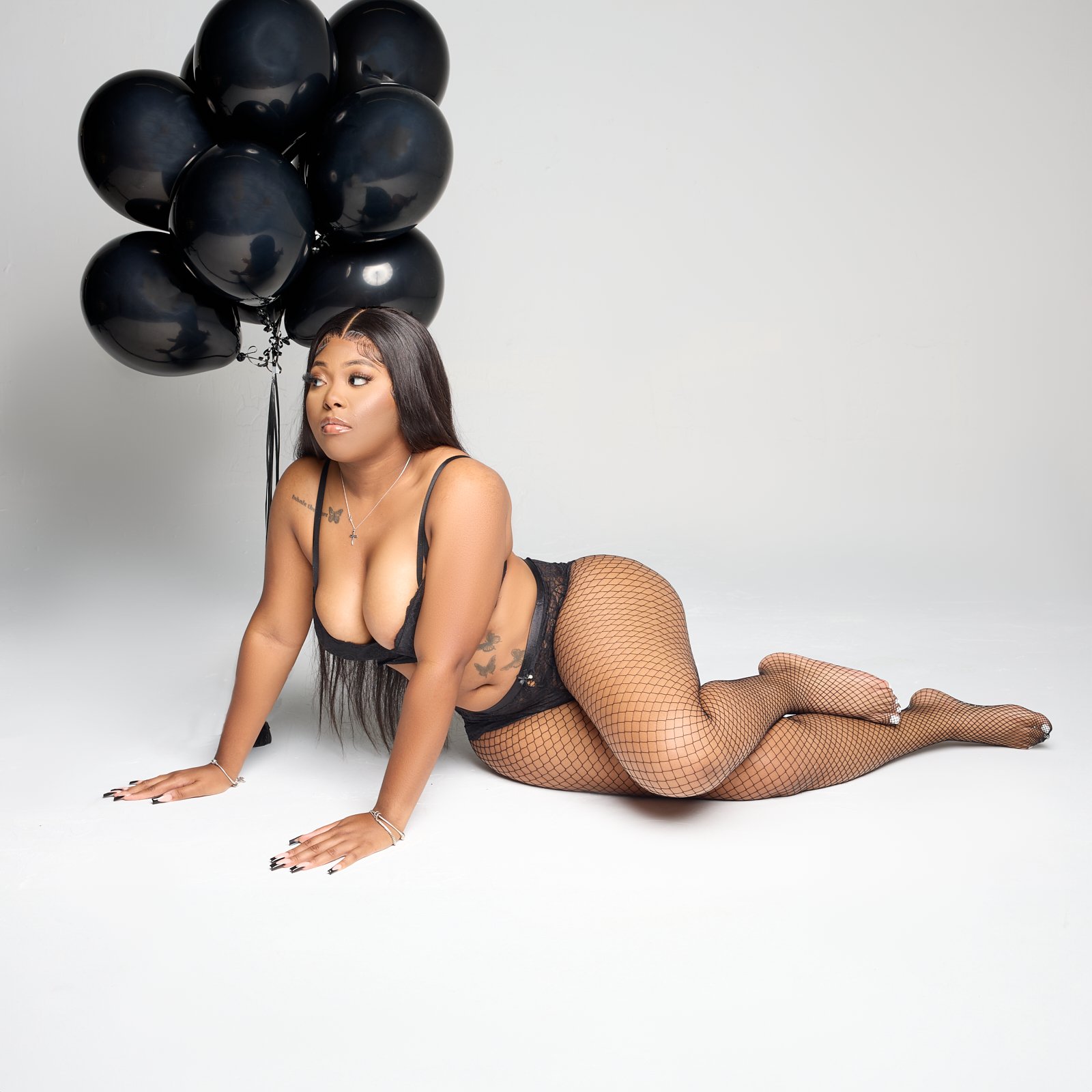 A woman lying on her hands and knees on the floor, wearing black lingerie and fishnet stockings, with black balloons attached to her, against a plain white background.