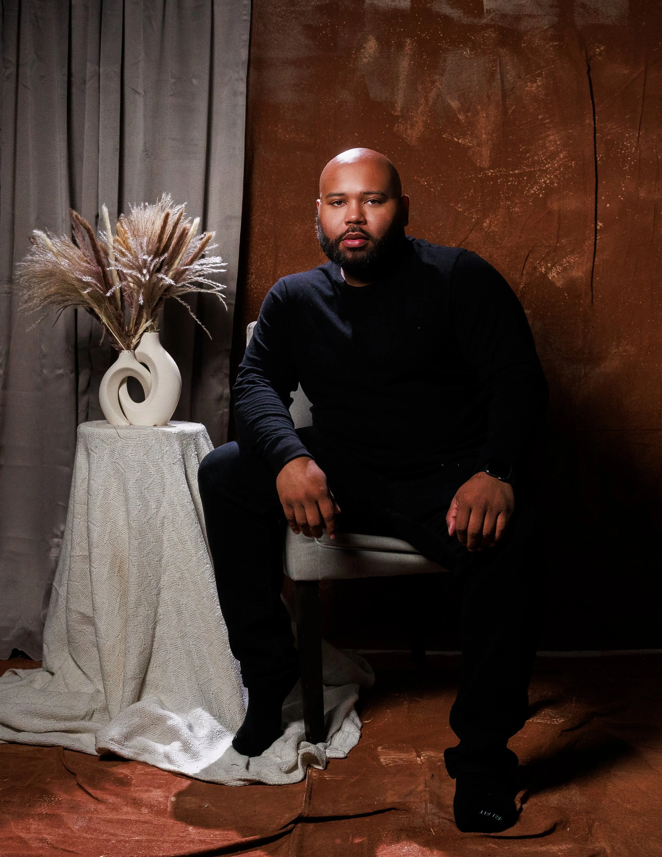 A bald man with a beard wearing a black long-sleeve shirt sitting on a chair against a textured brown wall. To his left, there is a white vase with dried pampas grass on a small table draped with a white cloth. The floor is wooden.