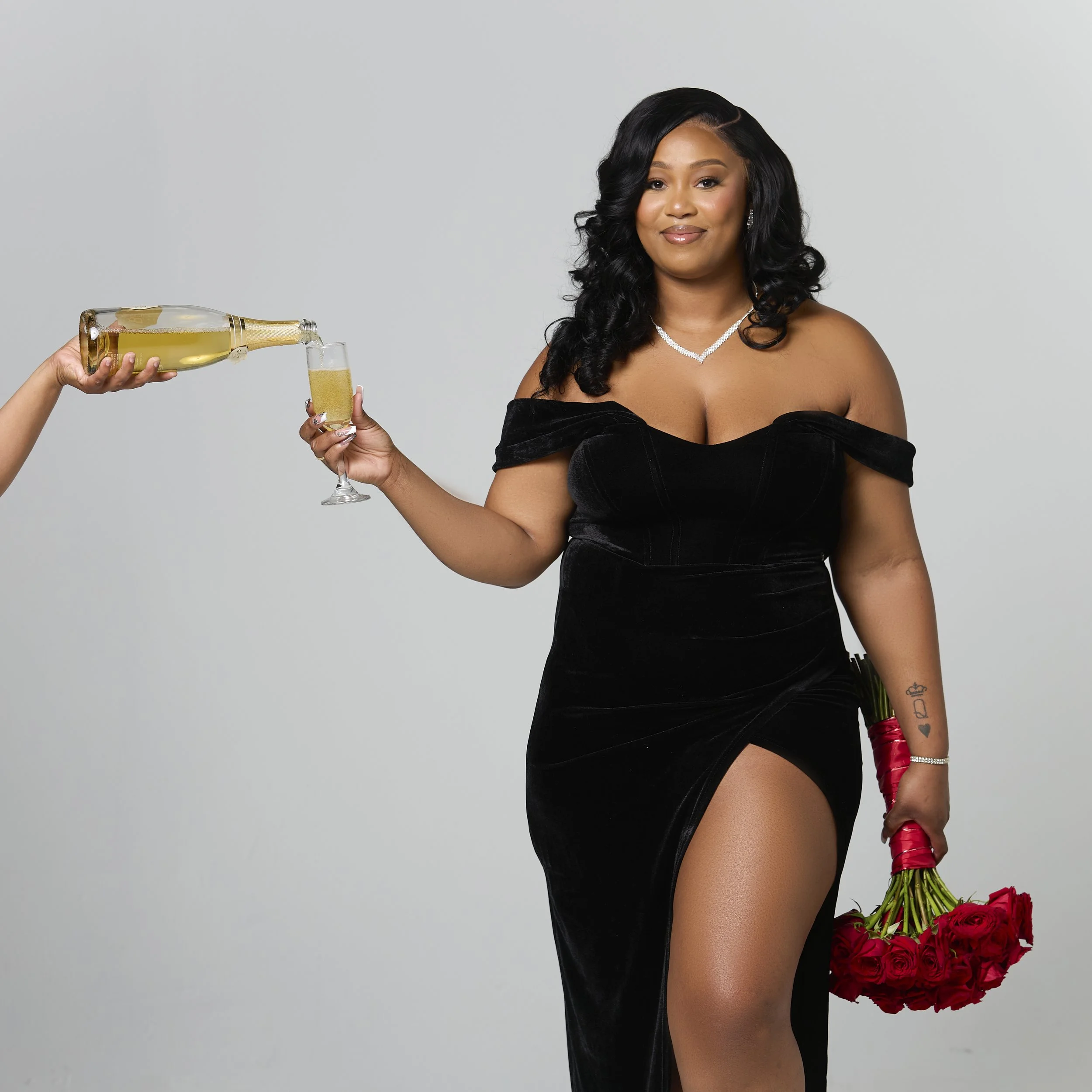A woman in a black dress holding a bouquet of red roses in one hand and a champagne bottle pouring into a glass held by someone else in the other hand, against a plain light background.