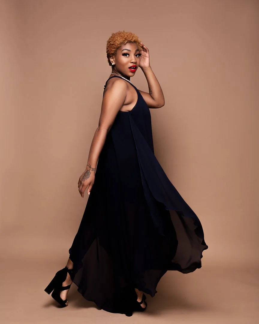 A woman with short, curly blonde hair wearing a black sleeveless dress and black high heels, posing against a beige background.