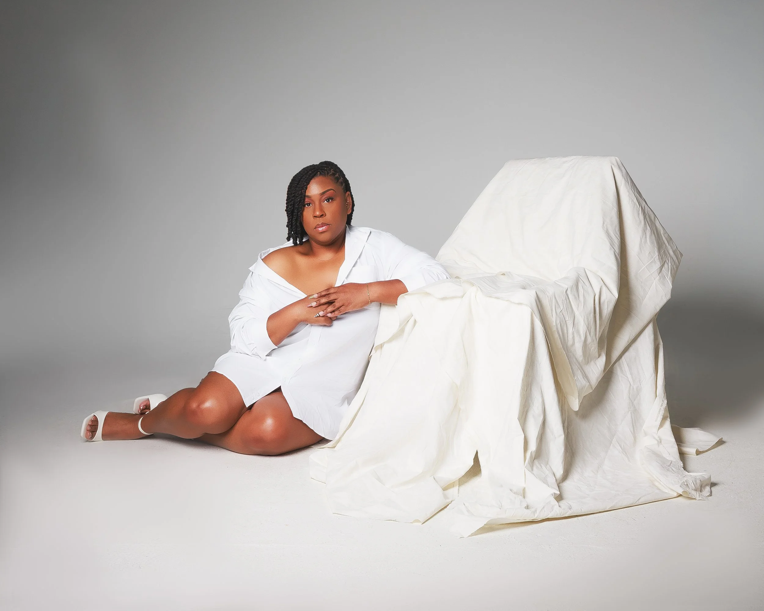 A woman with dark braided hair in a white blouse sitting on the floor with her legs crossed. She is leaning on an off-white, draped fabric or material, with a plain gray backdrop behind her.
